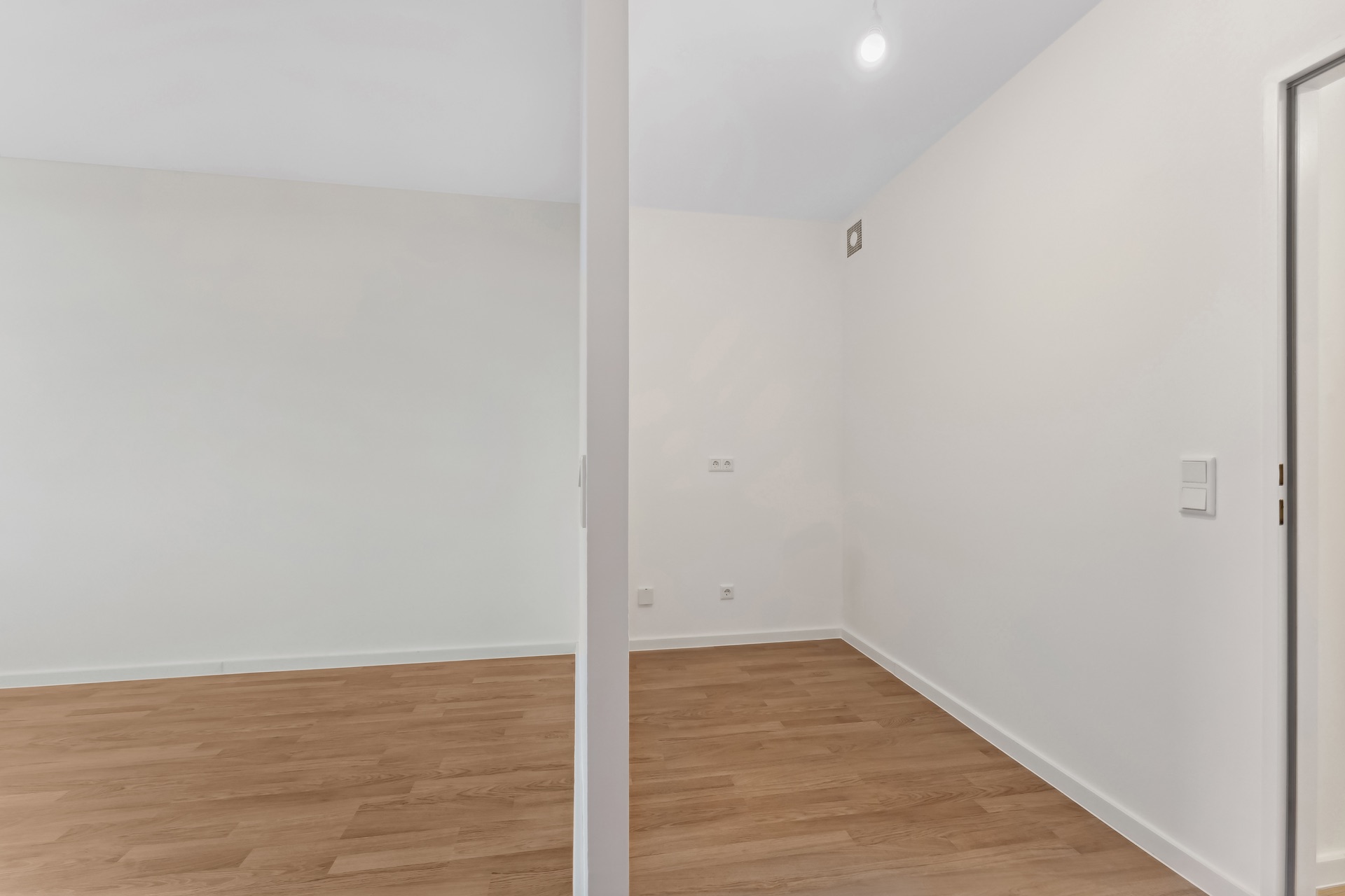 Zwei leere Zimmer mit weiß gestrichenen Wänden und Holzfußboden, getrennt durch eine Wand.