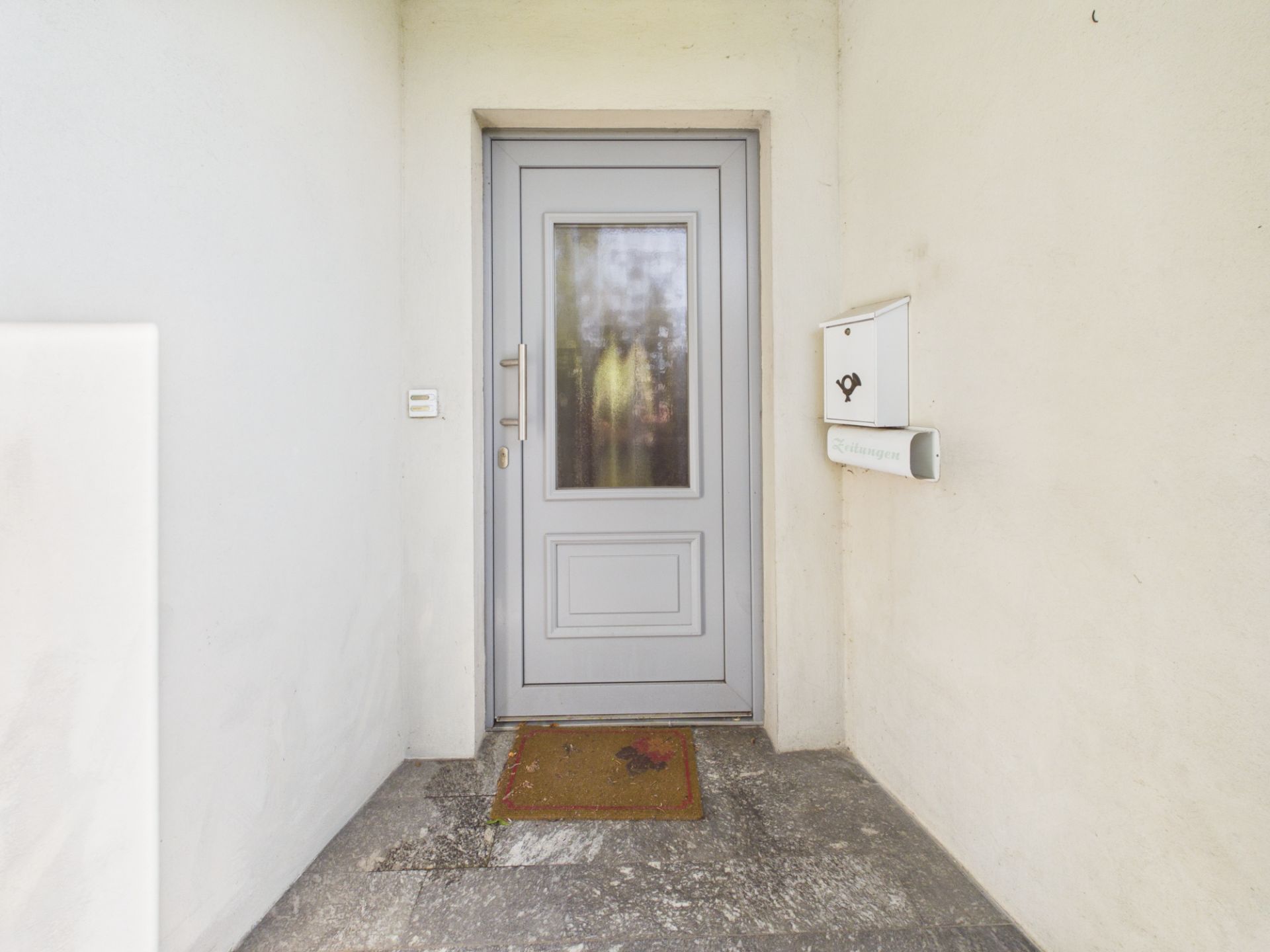 Eingangstür mit Glaseinsatz, Fußmatte und Briefkasten an einer hellen Hauswand.