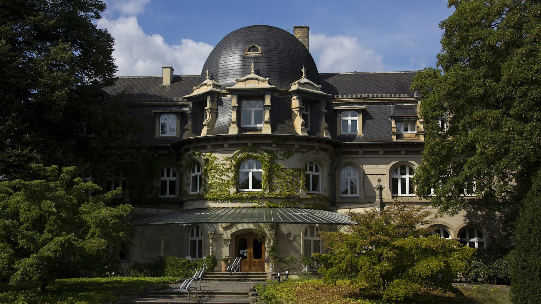 Großes Wohnhaus mit Kuppeldach und bewachsener Fassade, umgeben von Bäumen.