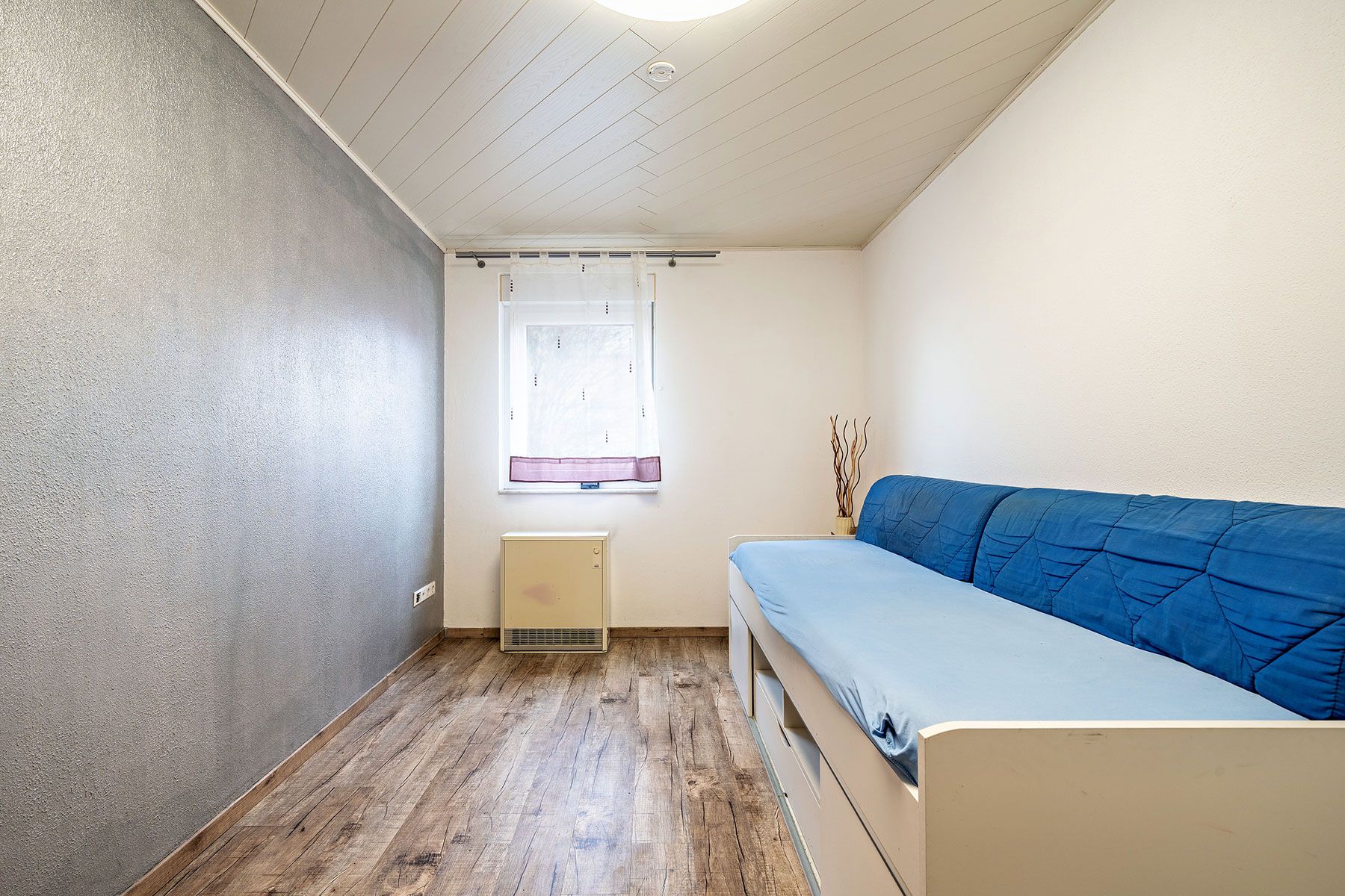 Kleines Zimmer mit Holzboden, einem Sofa mit blauem Bezug und einem Fenster mit Vorhang.