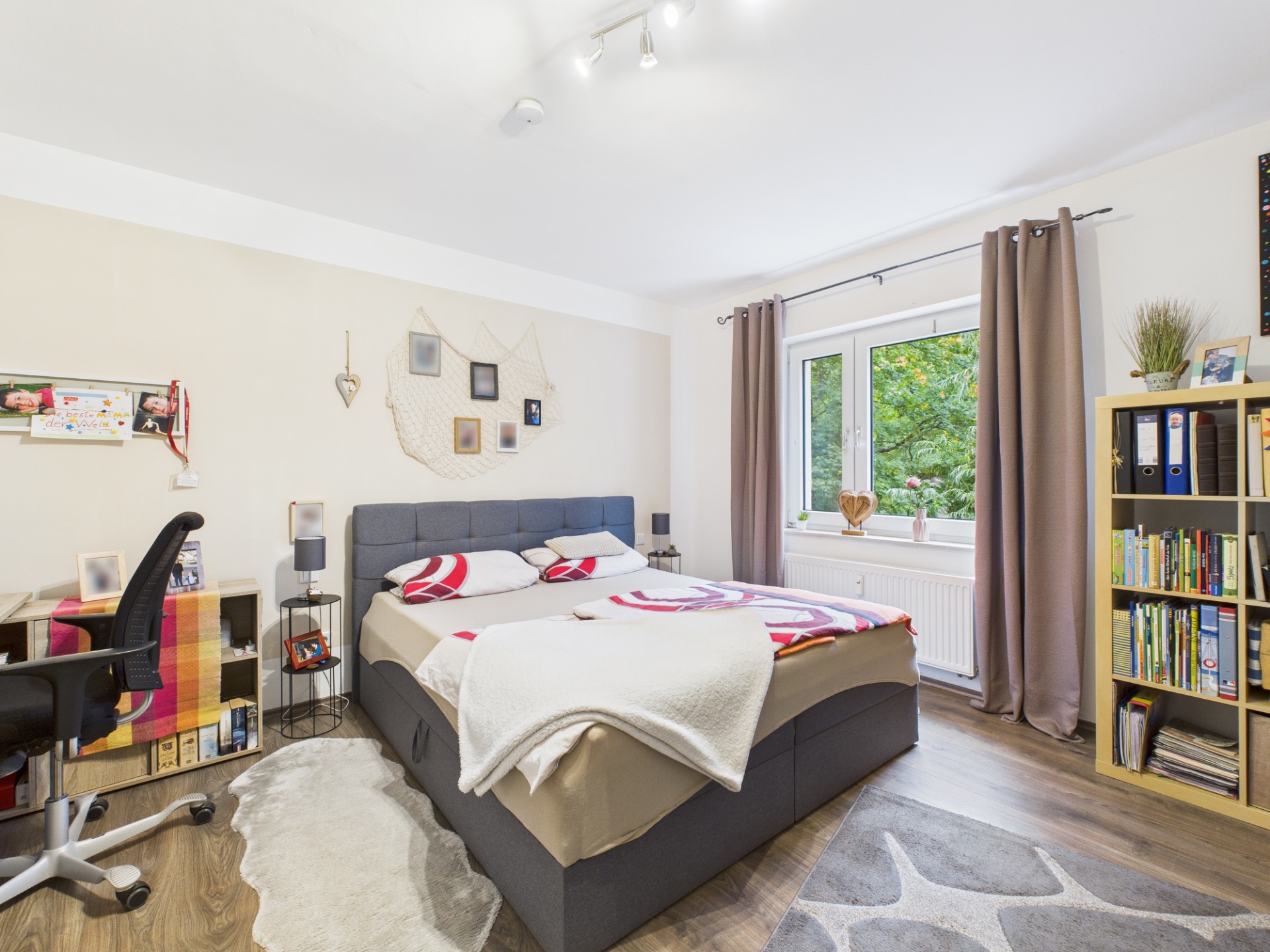 Helles Schlafzimmer mit Doppelbett, Schreibtisch, Bücherregal und großem Fenster mit Vorhängen.