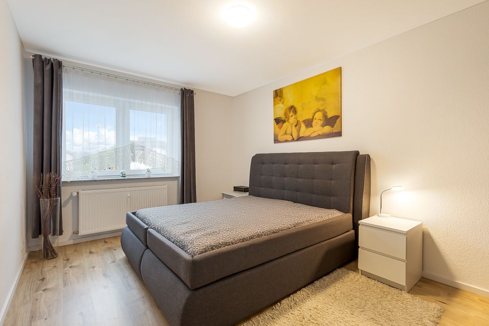 Modernes Schlafzimmer mit grauem Bett, Nachttisch, Lampe und großem Fenster mit Vorhängen.