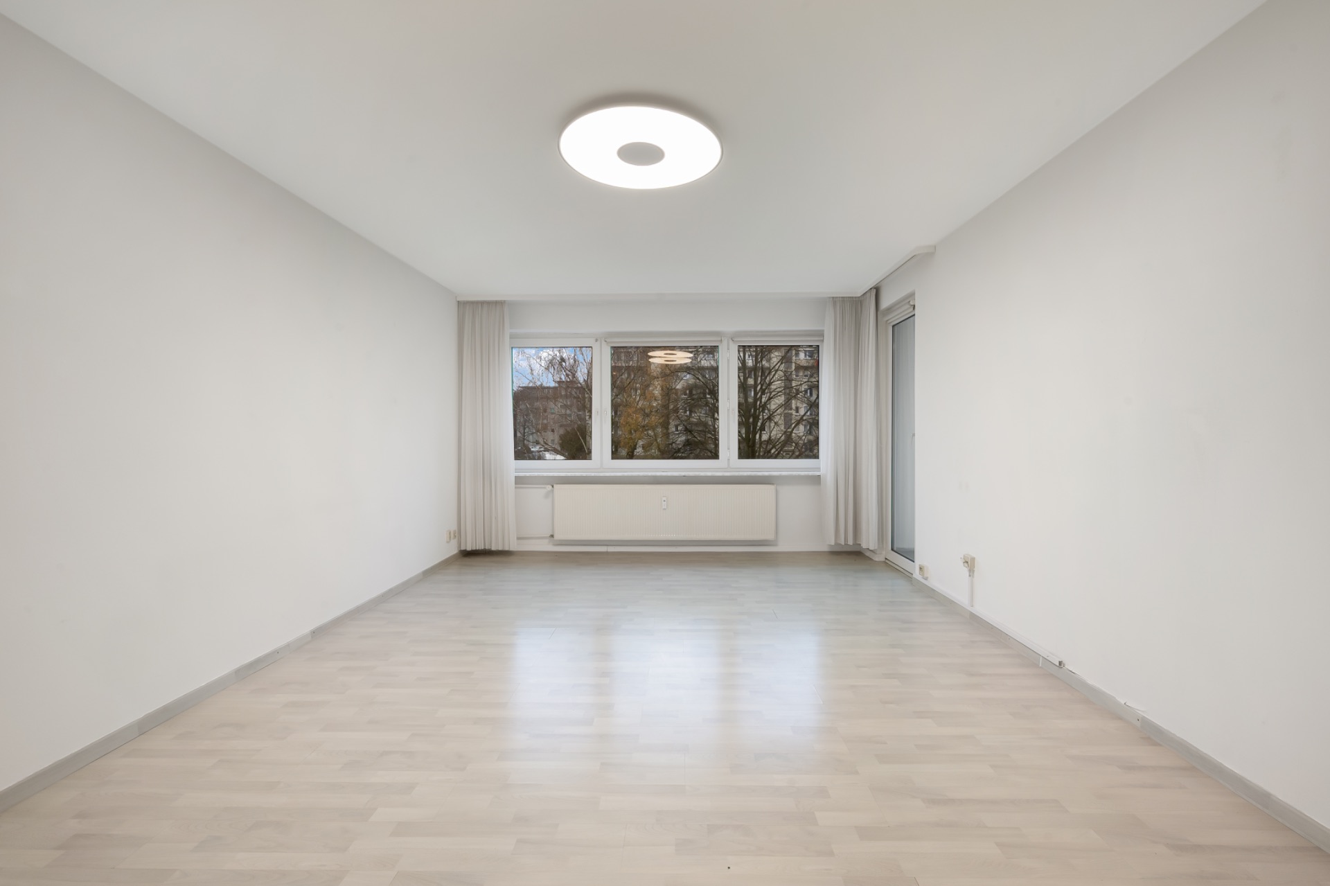 Helles, leeres Wohnzimmer mit großen Fenstern, hellem Boden und Deckenleuchte.