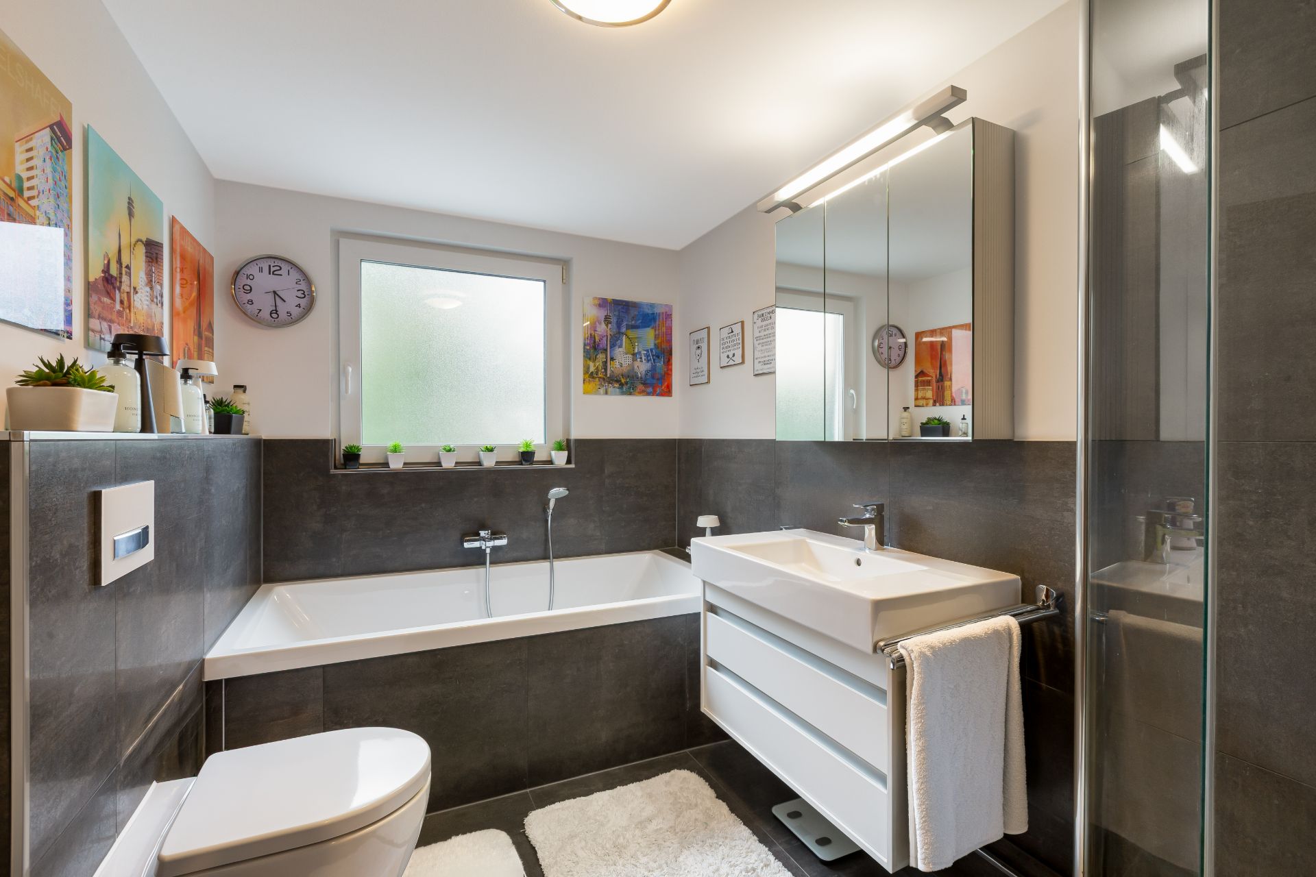 Modernes Badezimmer mit Badewanne, Dusche, Waschbecken und Spiegel, dekoriert mit Pflanzen und Bildern.