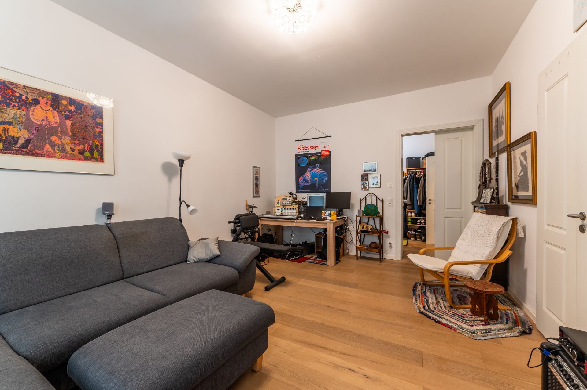 Wohnzimmer mit grauem Sofa, Schreibtisch, Stuhl, Bildern an der Wand und Holzboden.