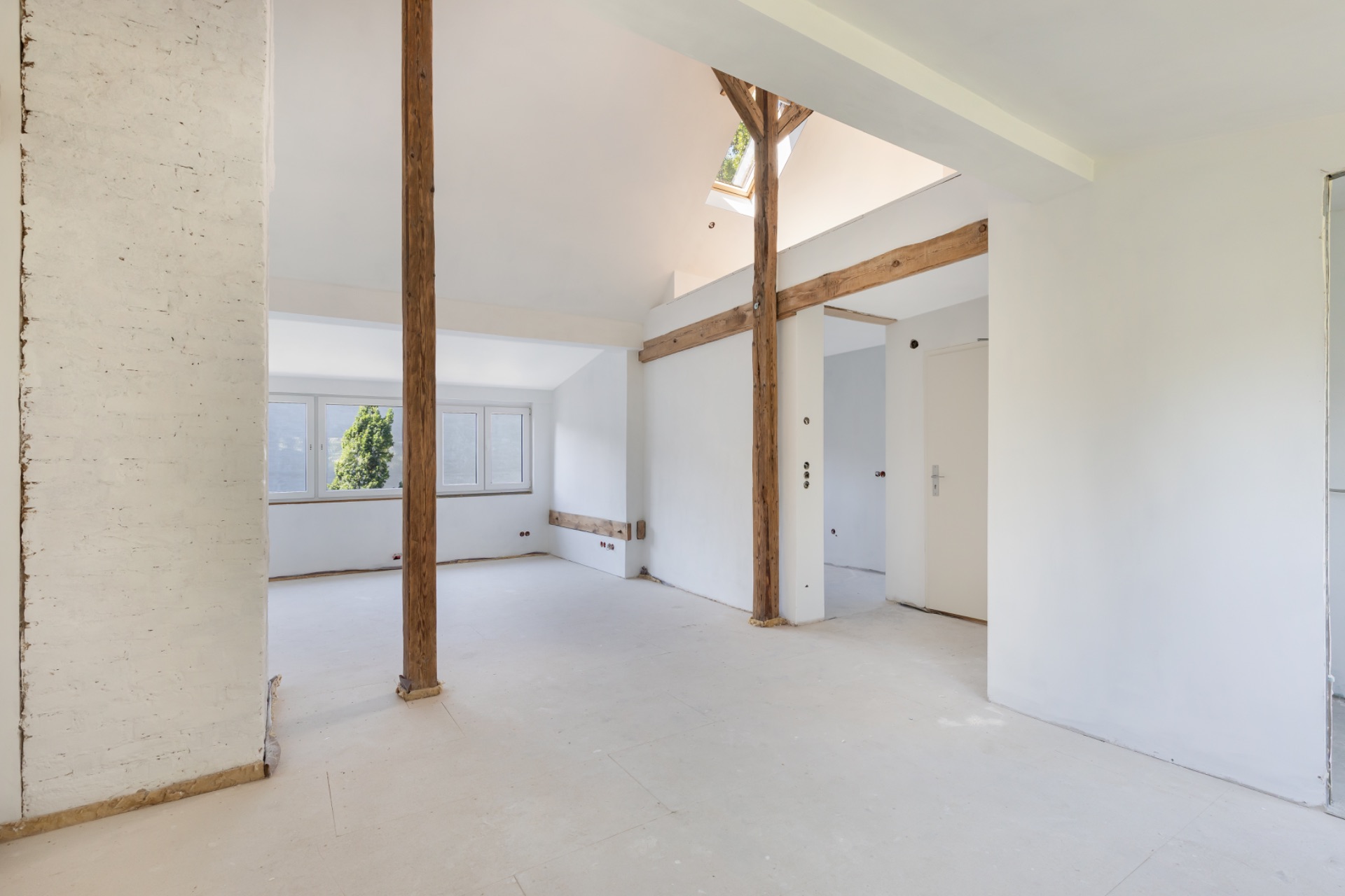 Heller, leerer Raum mit weißen Wänden, Holzbalken und großen Fenstern.