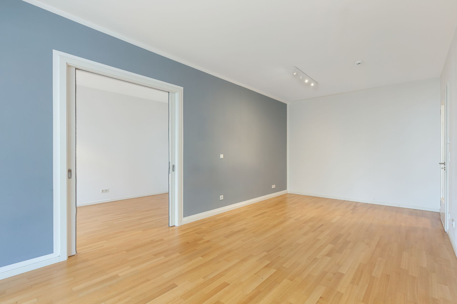 Heller, leerer Raum mit Holzfußboden und blauer Wand, Schiebetür zu weiterem Zimmer.