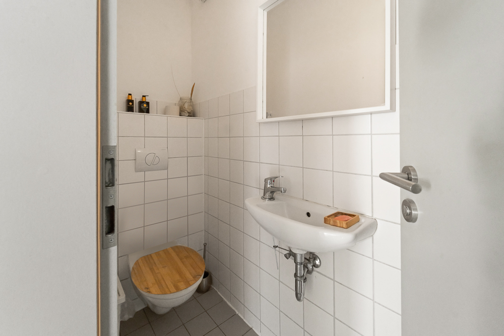 Kleines Badezimmer mit Wandtoilette, Waschbecken, Spiegel und weiß gefliesten Wänden.