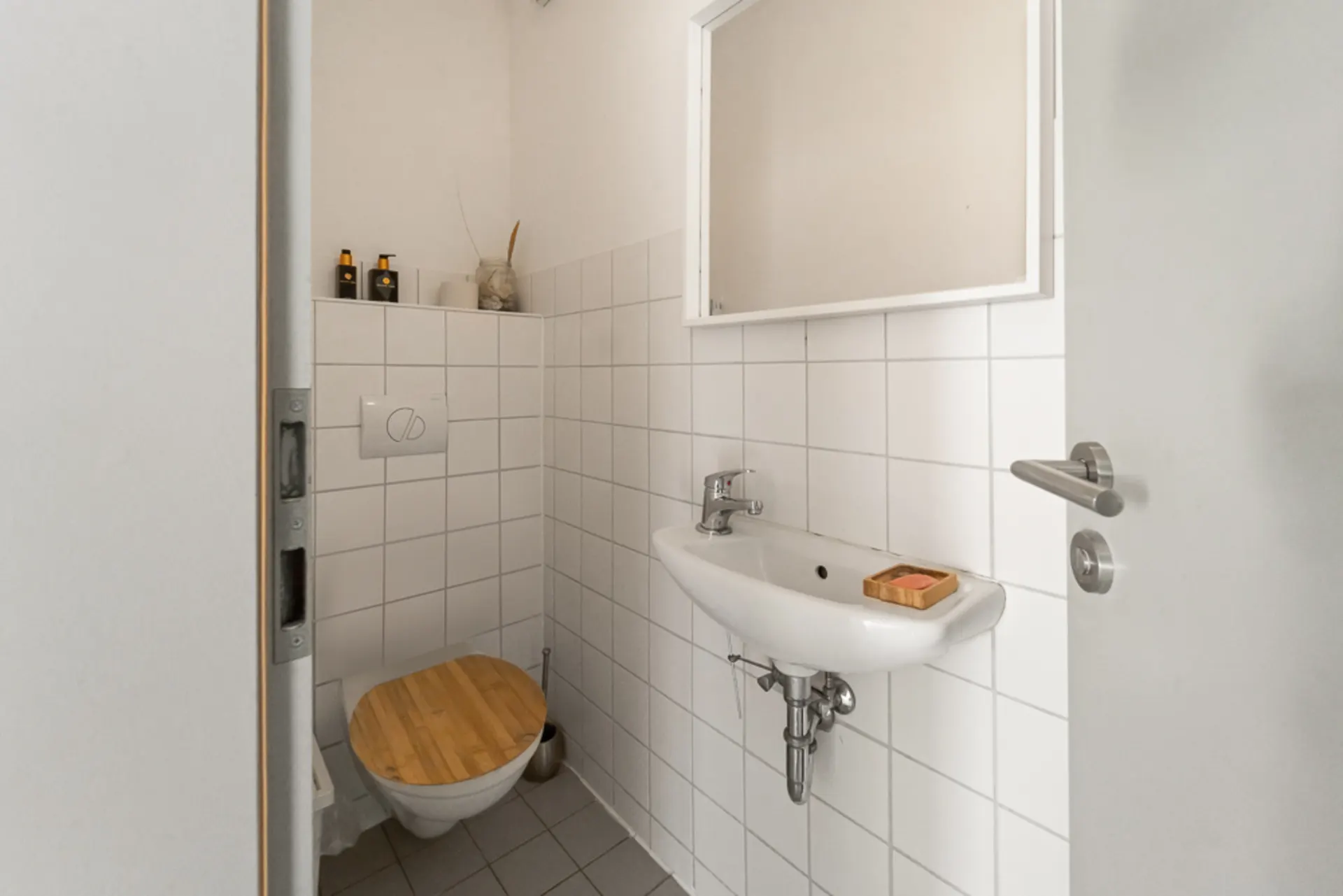 Kleines Badezimmer mit Wandtoilette, Waschbecken, Spiegel und weiß gefliesten Wänden.