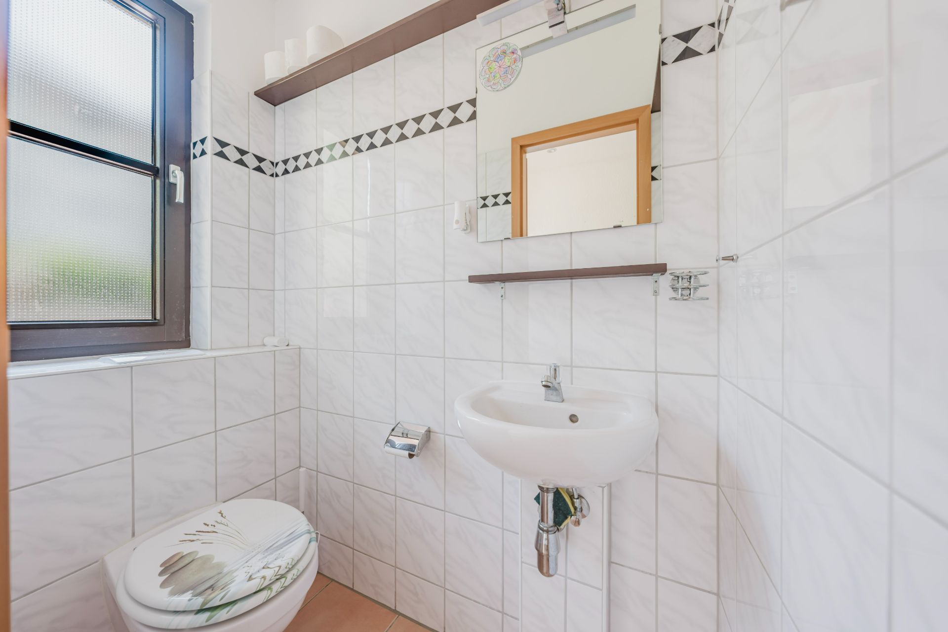 Kleines Badezimmer mit Fenster, Toilette, Waschbecken und Spiegel, weiße Fliesen an den Wänden.