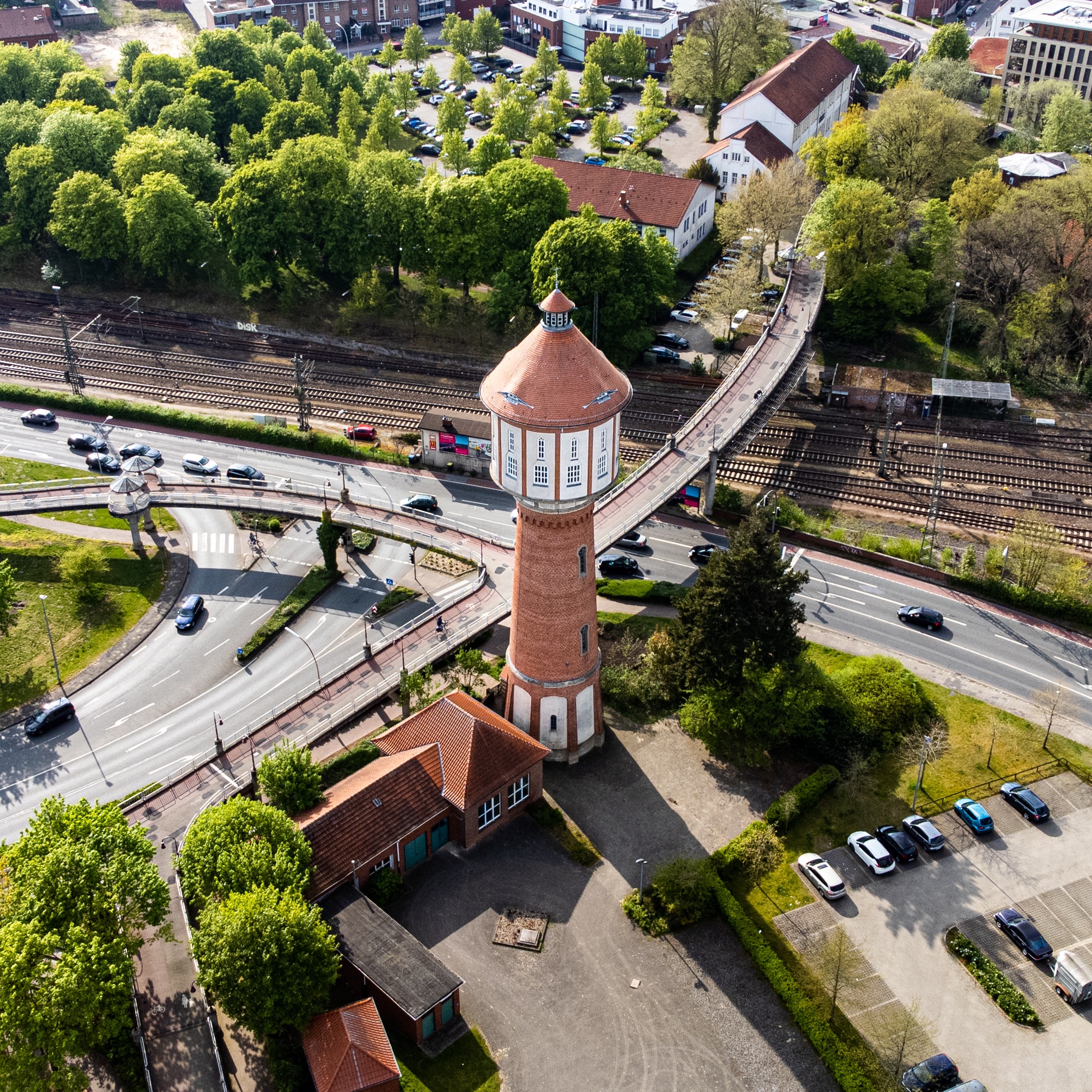 Luftaufnahme eines roten Wasserturms neben Bahnlinien und Straßen, umgeben von Bäumen und Parkplätzen.