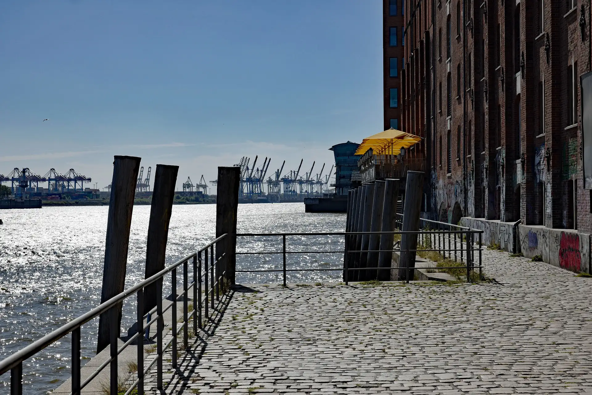 Kopfsteinpflasterweg am Wasser neben einem Backsteingebäude mit gelben Sonnenschirmen.