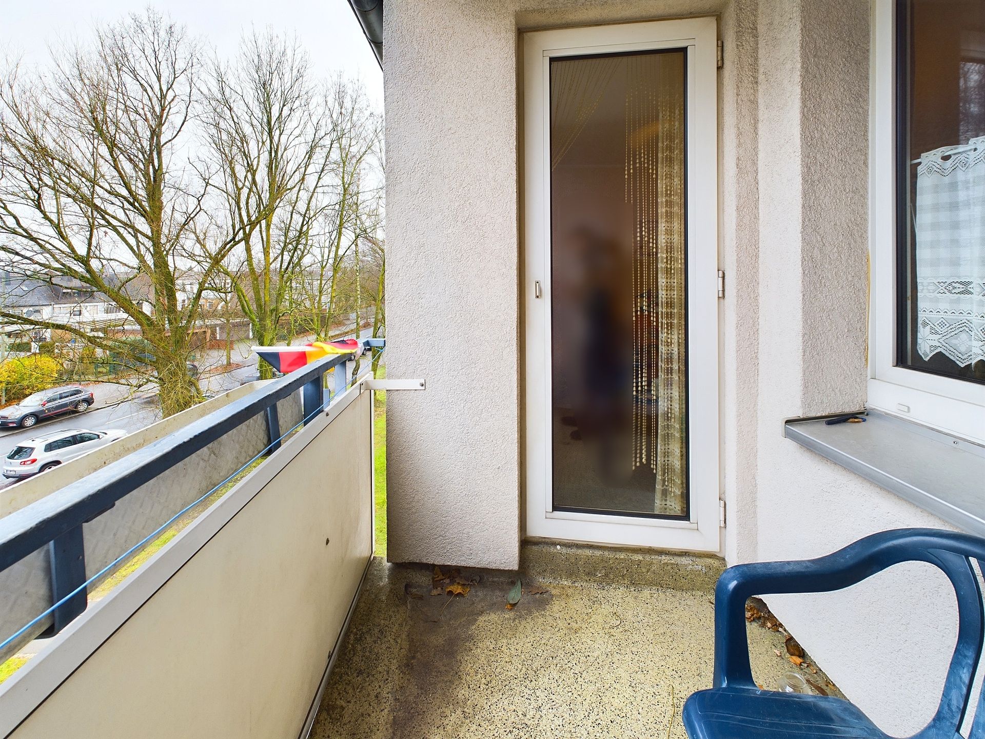Kleiner Balkon mit Kunststoffstuhl und Blick auf Bäume und parkende Autos.