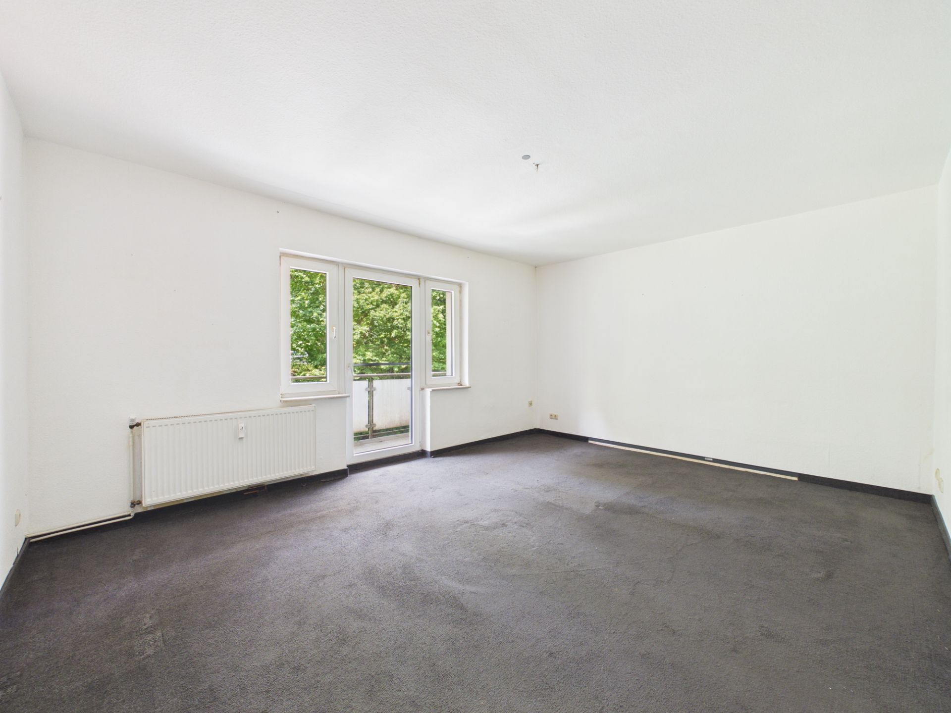 Leerer Raum mit weißer Wand, dunklem Teppich und Fenster mit Blick auf Bäume.