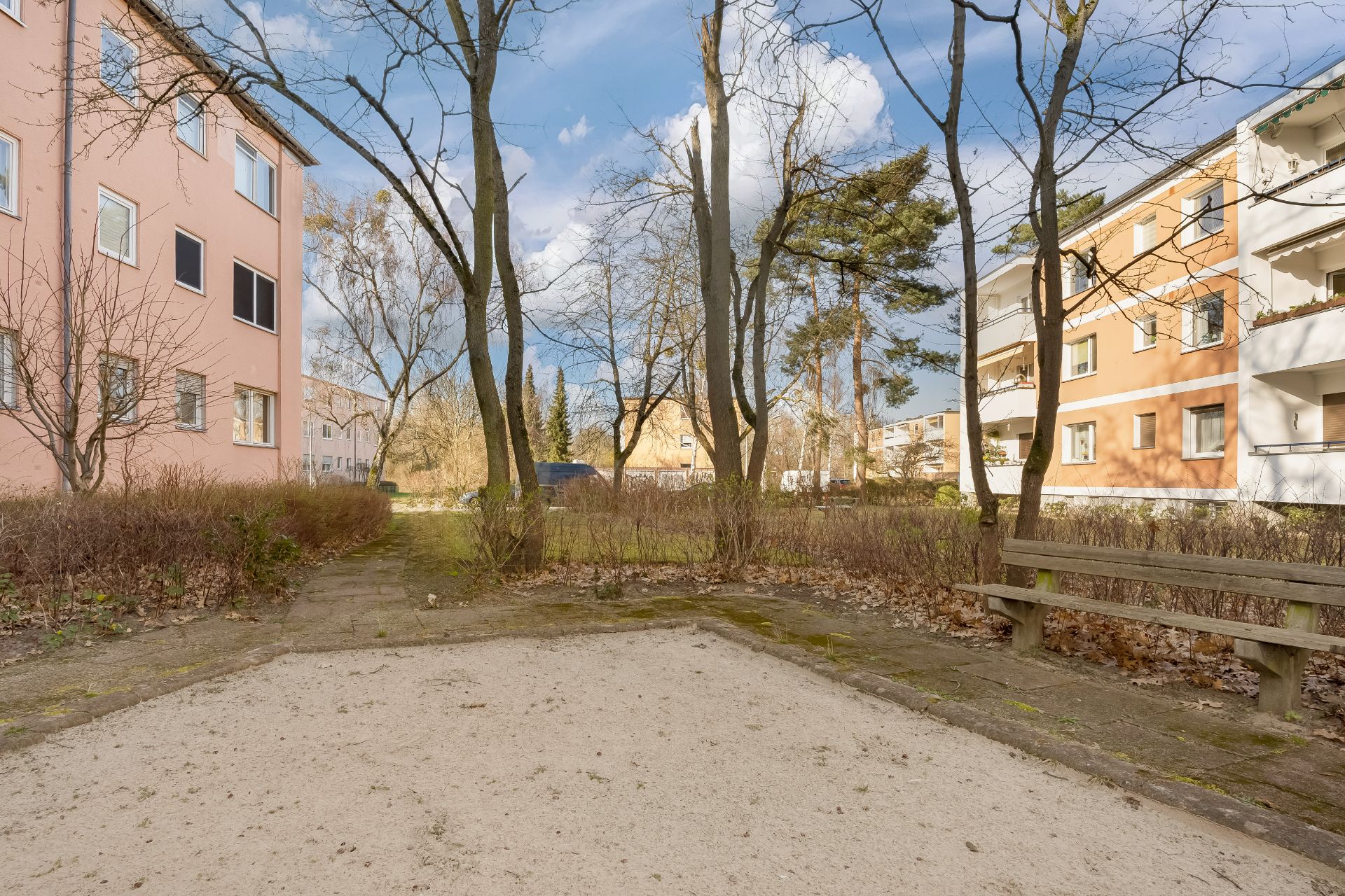 Wohnhof mit Bäumen, Sandfläche, Bank und angrenzenden Mehrfamilienhäusern bei Tageslicht.
