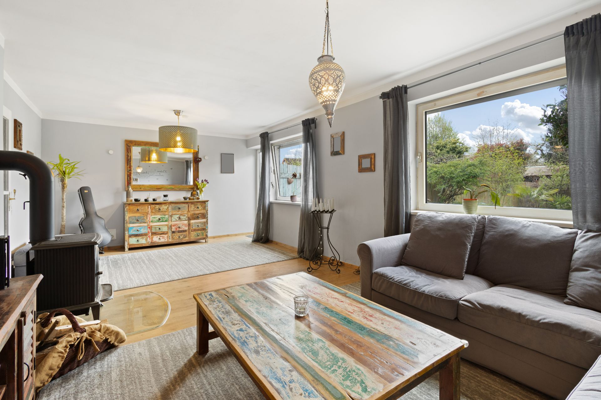 Helles Wohnzimmer mit grauem Sofa, Couchtisch aus Holz und zwei großen Fenstern mit Blick ins Grüne.