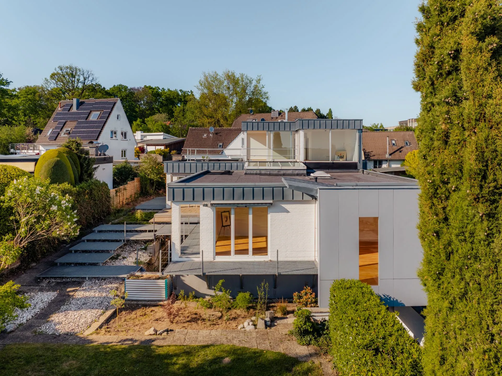 Modernes Wohnhaus mit flachem Dach, großen Fenstern und Garten, umgeben von weiteren Häusern.