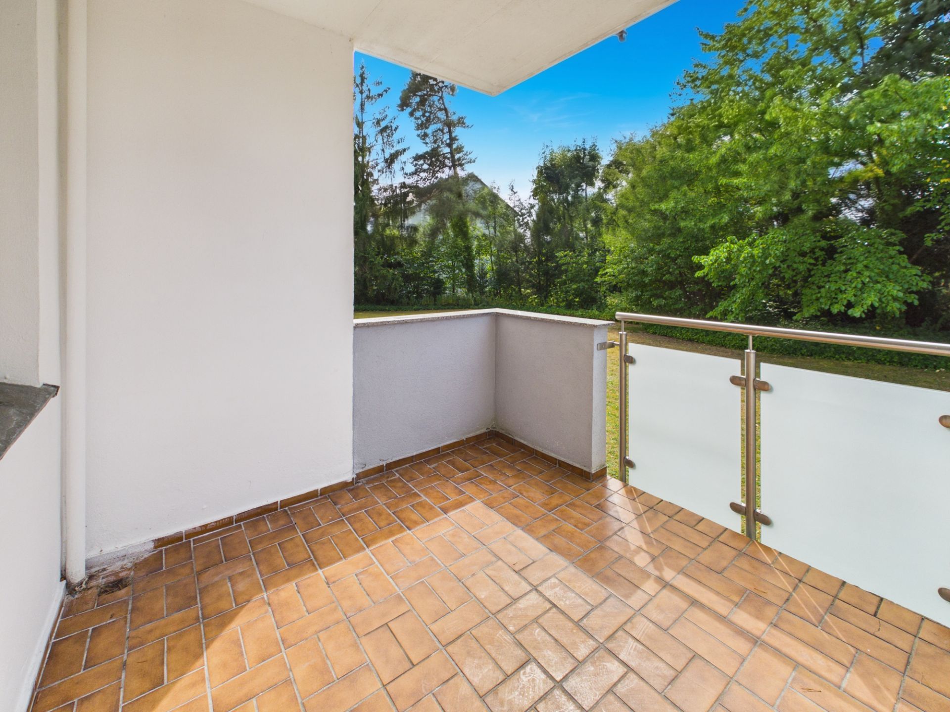 Balkon mit braunen Fliesen, Glasgeländer und Blick auf grüne Bäume.