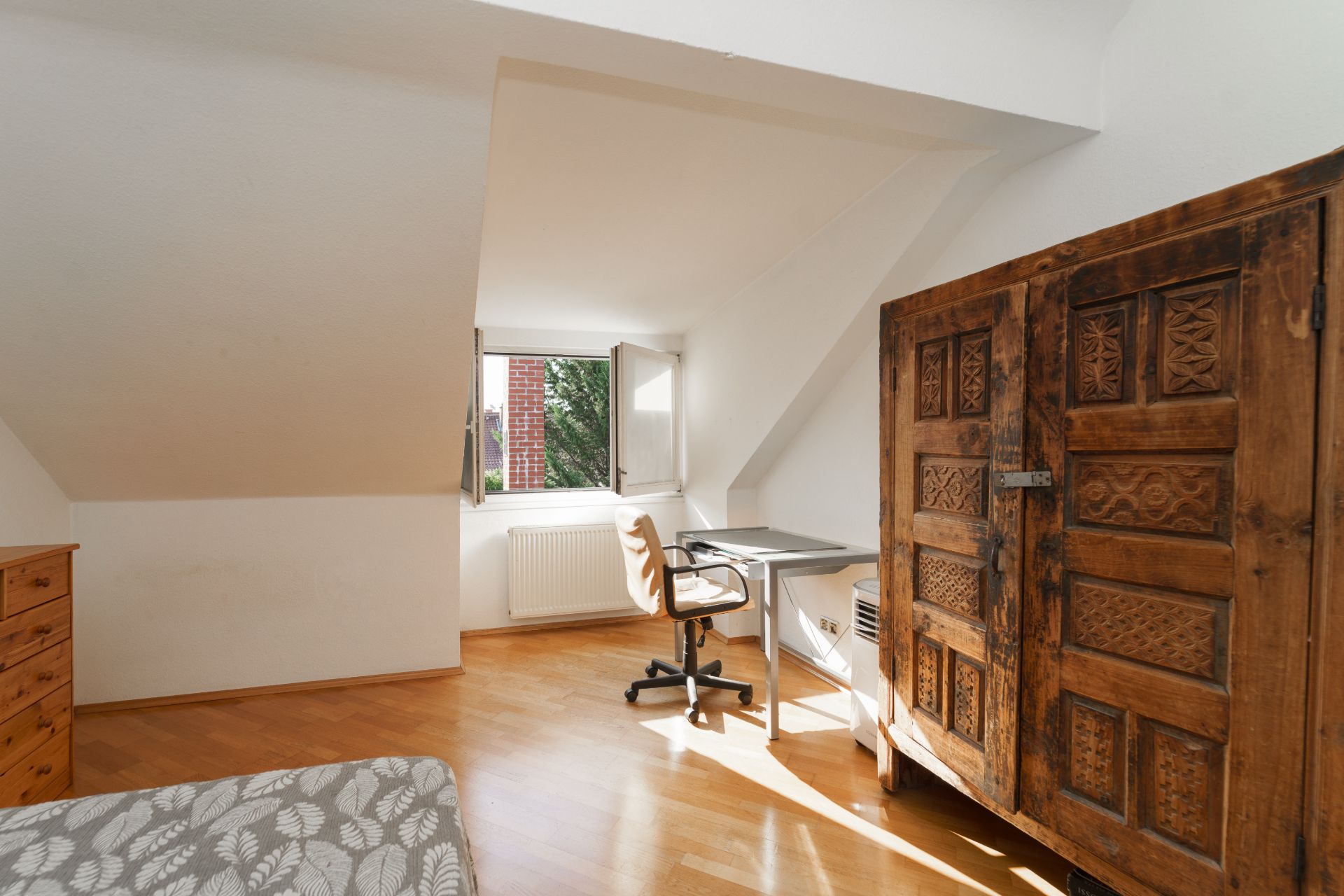 Helles Zimmer mit Holzschrank, Schreibtisch am Fenster und Stuhl auf Parkettboden.
