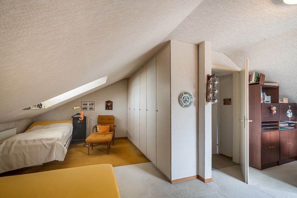 Dachzimmer mit Bett, Sessel, Einbauschränken und Regal, Tageslicht durch Dachfenster.