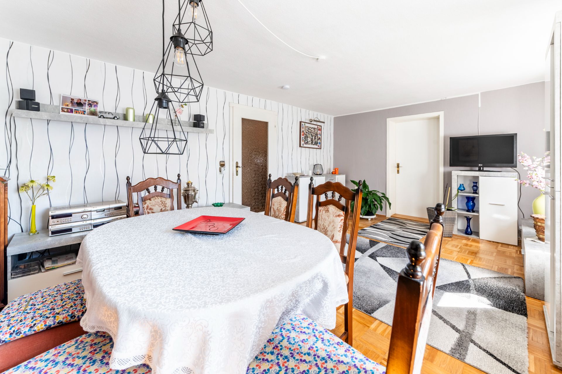 Helles Wohnzimmer mit rundem Esstisch, Stühlen, Regal, Teppich und Fernseher.
