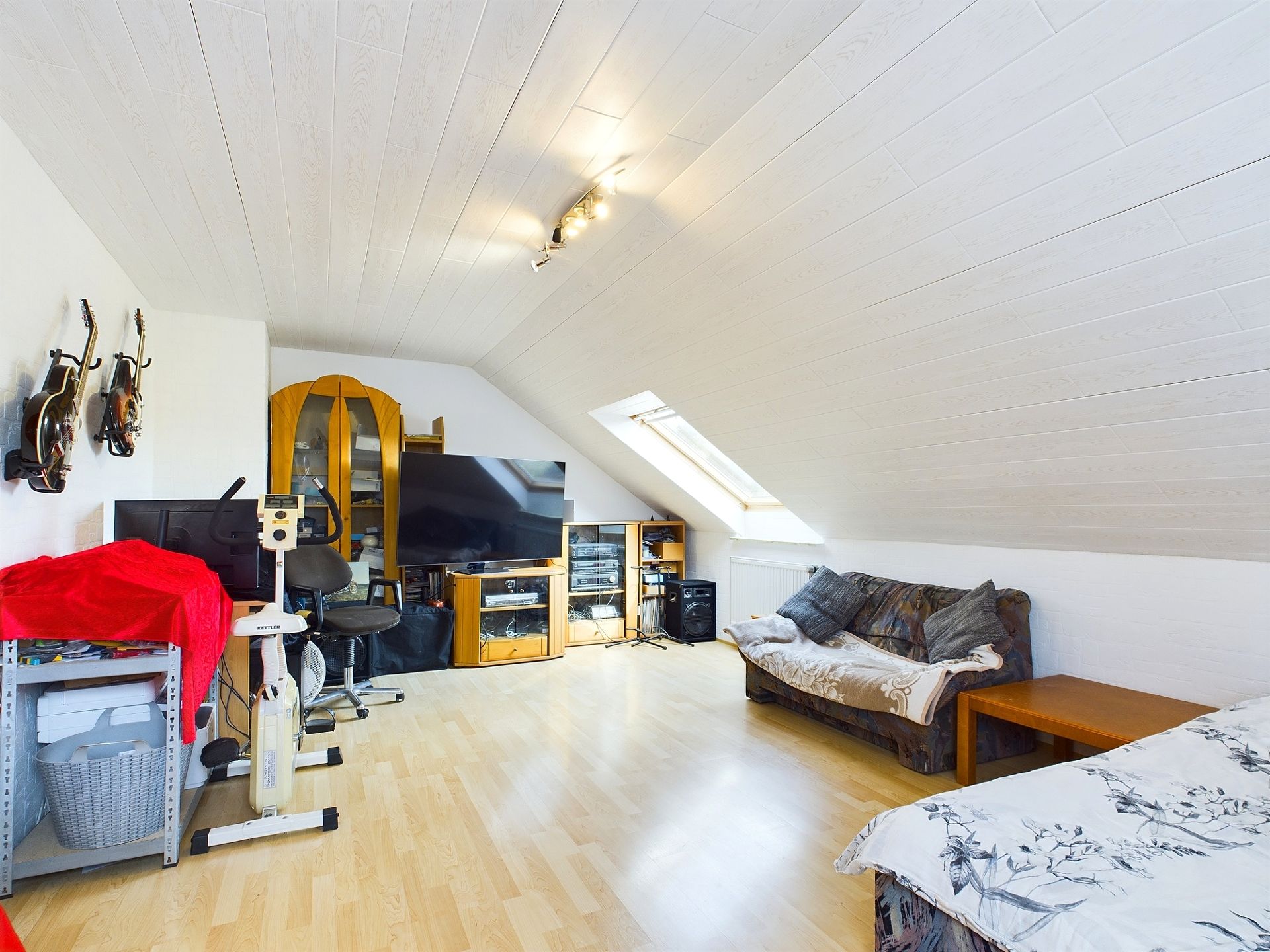Helles Dachzimmer mit Sofa, Fernseher, Schreibtisch und Musikinstrumenten an der Wand.