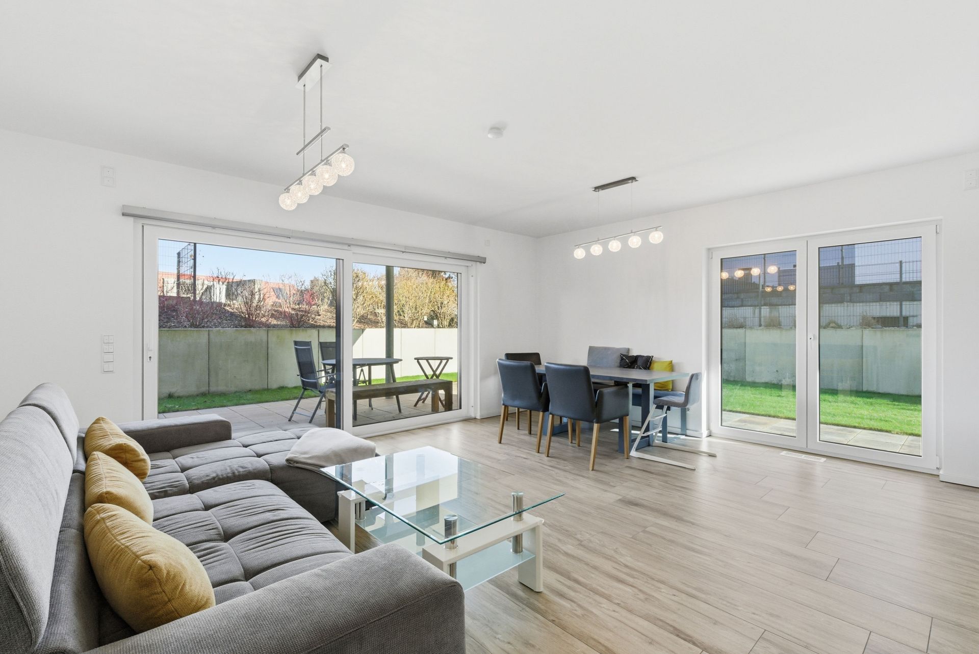 Modernes Wohnzimmer mit grauem Sofa, Glastisch, Esstisch, großen Fenstern und Blick auf den Garten.