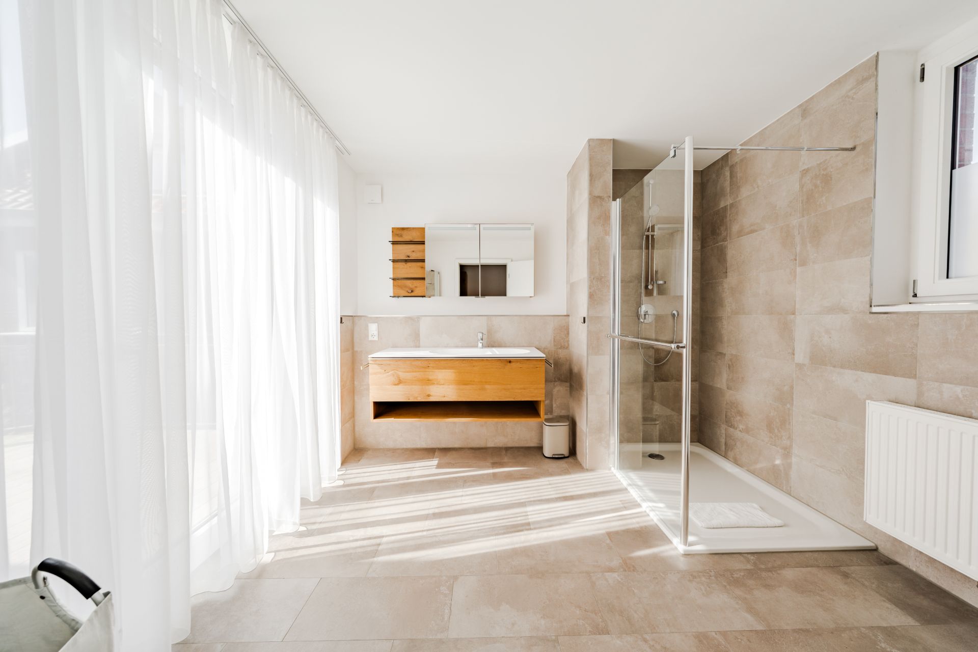 Modernes Badezimmer mit Glasdusche, Holzwaschtisch und großem Fenster mit weißen Vorhängen.