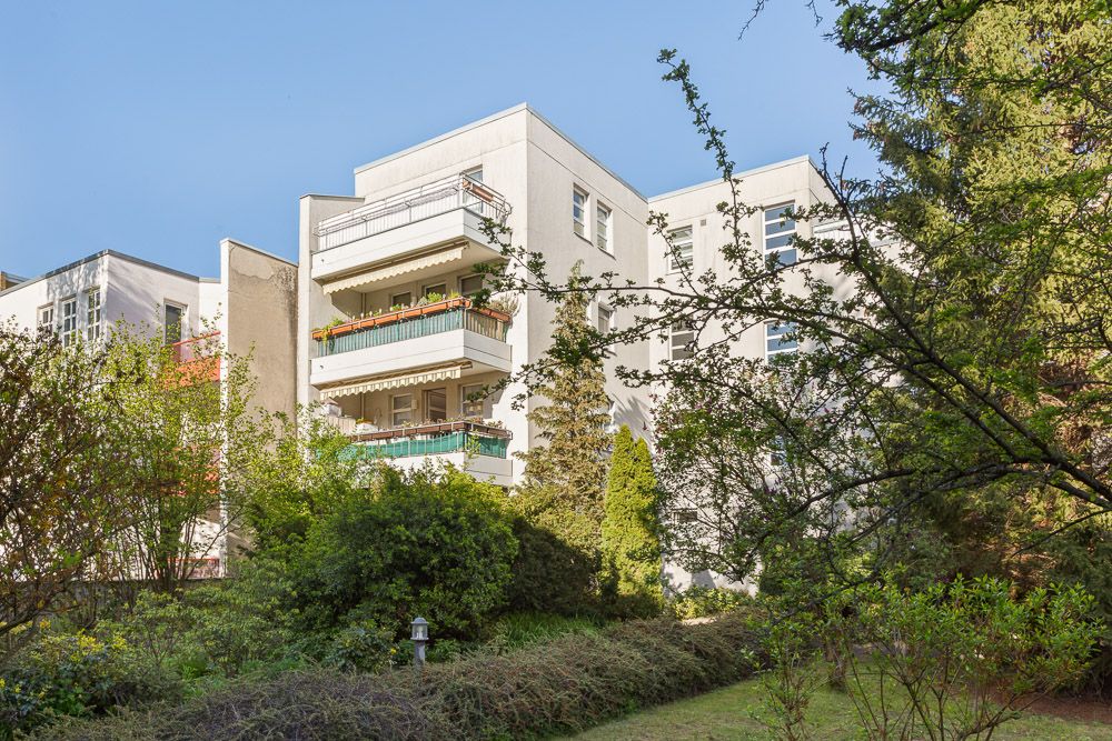 Mehrstöckiges Wohnhaus mit Balkonen, umgeben von Bäumen und Garten im Vordergrund.