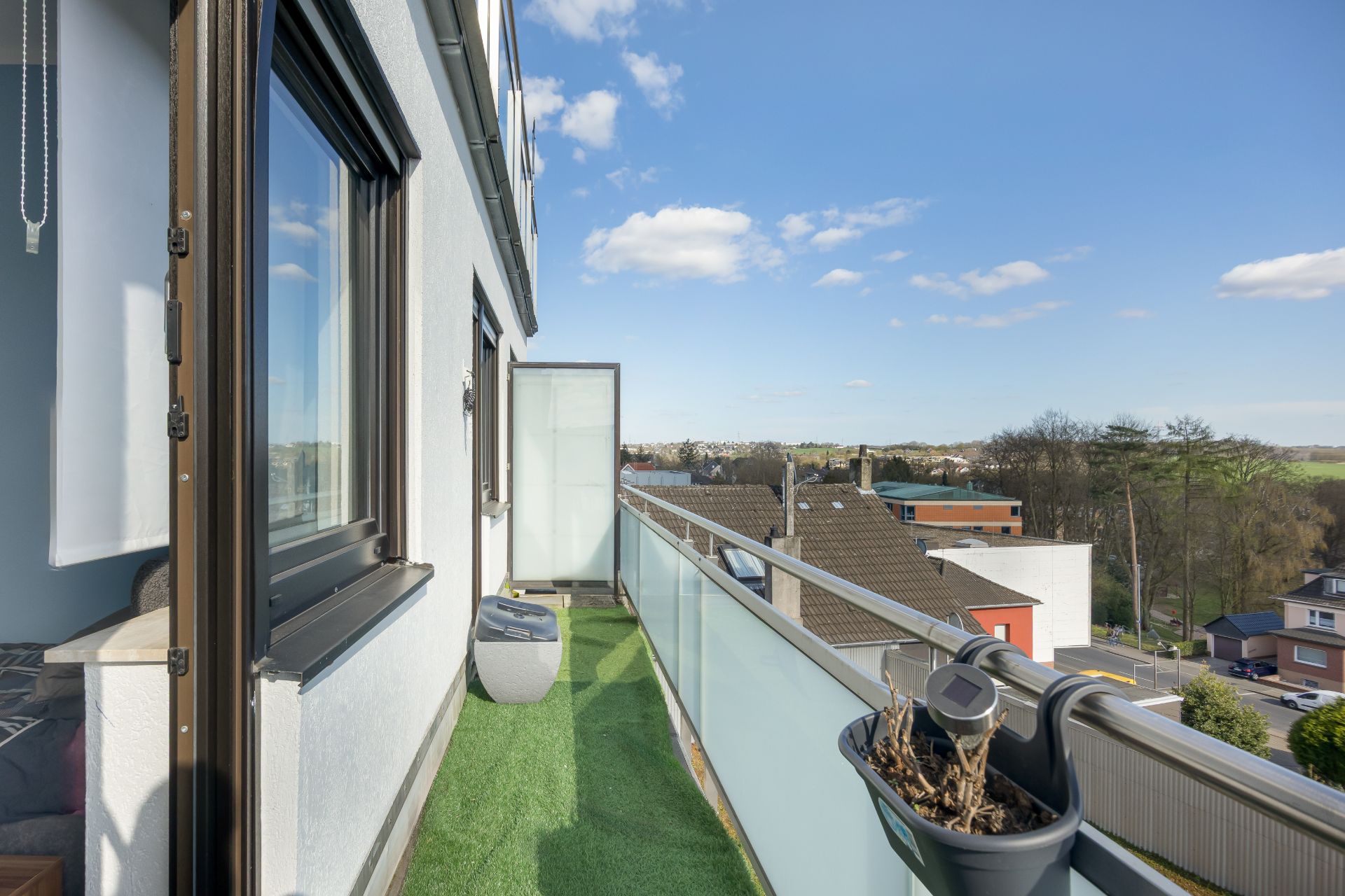 Schmaler Balkon mit Kunstrasen und Pflanzgefäß, Blick auf Nachbarhäuser und blauen Himmel.