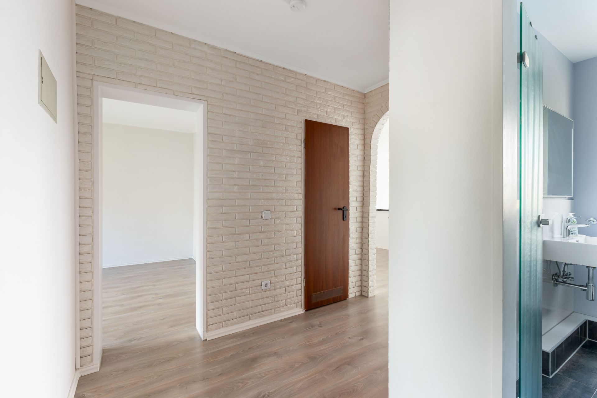 Heller Flur mit Holzboden, weißer Ziegelwand, Holztür und Sicht ins Badezimmer mit Waschbecken.