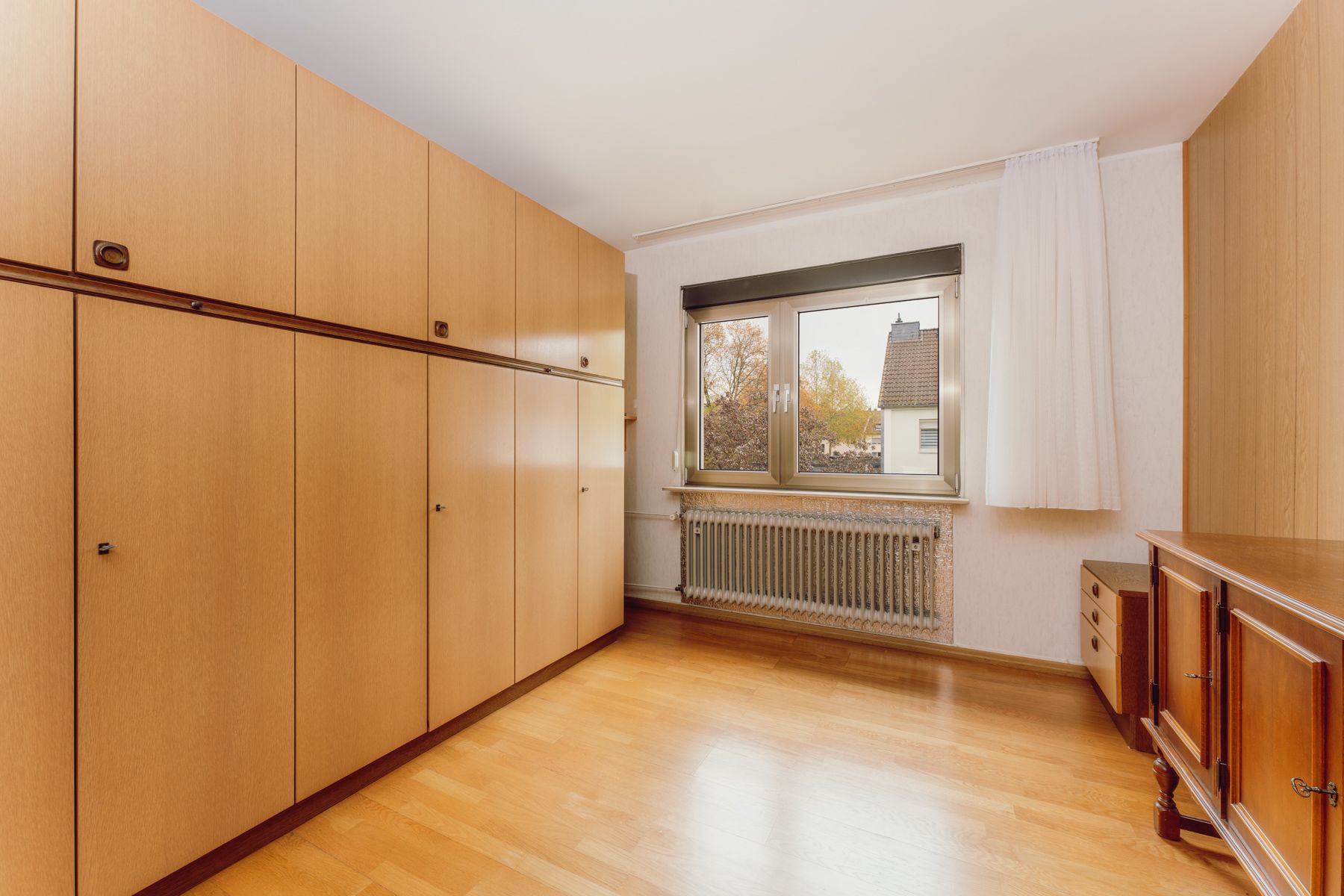 Helles Zimmer mit großen Holzschränken, Fenster, Heizkörper und Parkettboden.