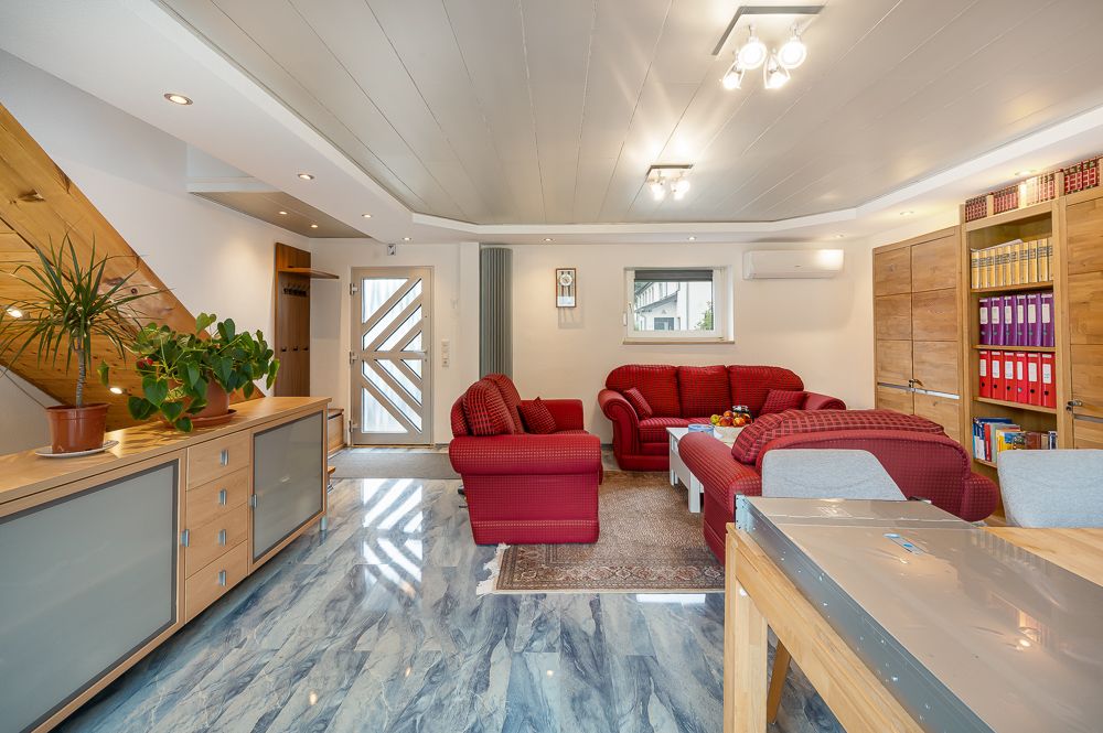 Wohnzimmer mit roten Sofas, Holzmöbeln, Pflanzen und Regalen mit Ordnern auf blauem Marmorboden.