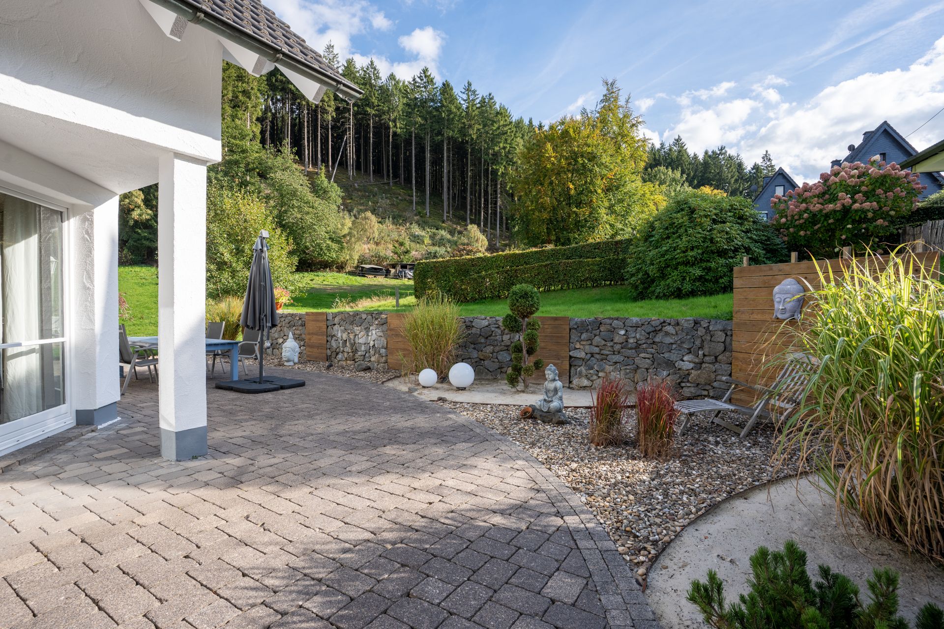 Gepflasterte Terrasse mit Garten, Pflanzen, Gartenmöbeln und Blick auf einen bewaldeten Hang.