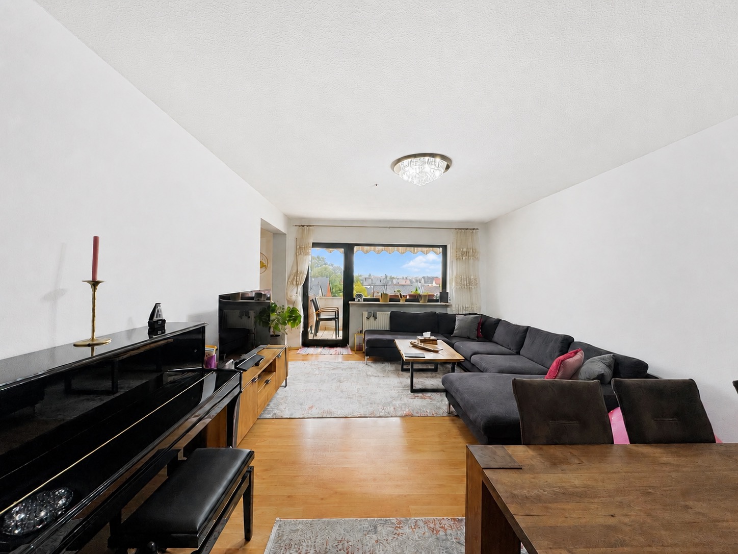 Helles Wohnzimmer mit großem Ecksofa, Klavier und Esstisch, Blick auf Fenster mit Aussicht.