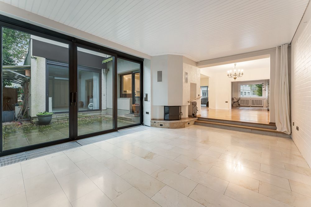 Geräumiges Wohnzimmer mit Kamin, großen Fenstern und Zugang zur Terrasse, daneben ein erhöhter Bereich mit Kronleuchter.