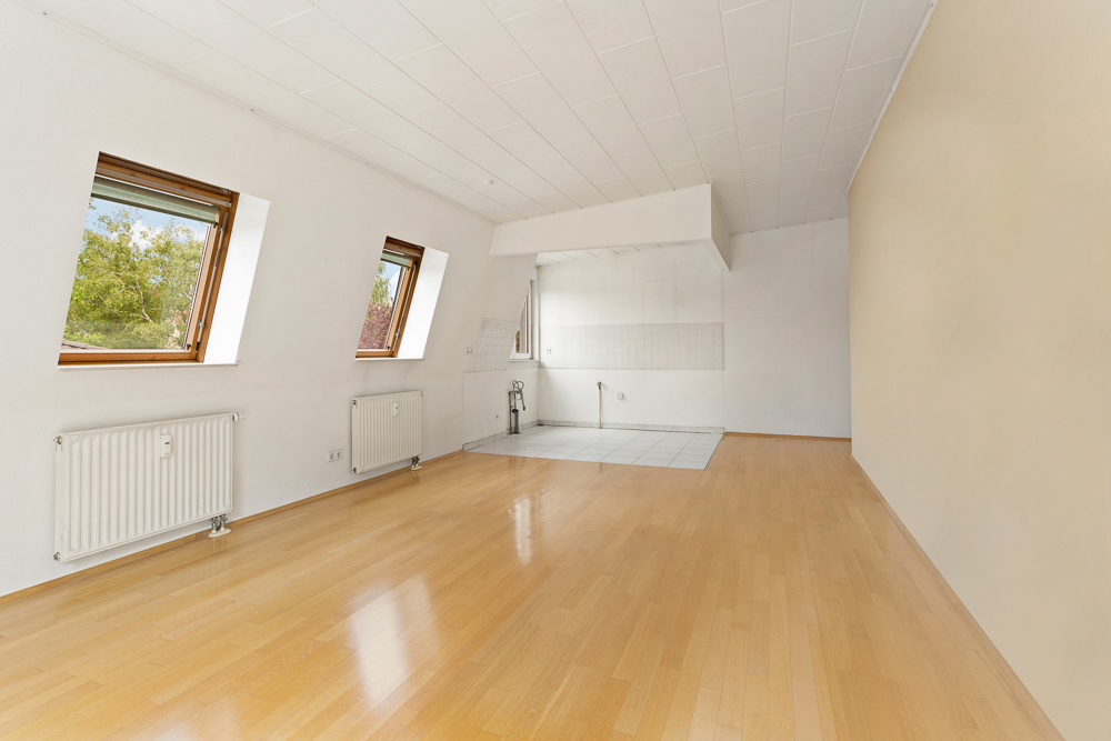 Leerer Raum mit zwei Fenstern, Holzboden und gefliestem Bereich für eine Küche.