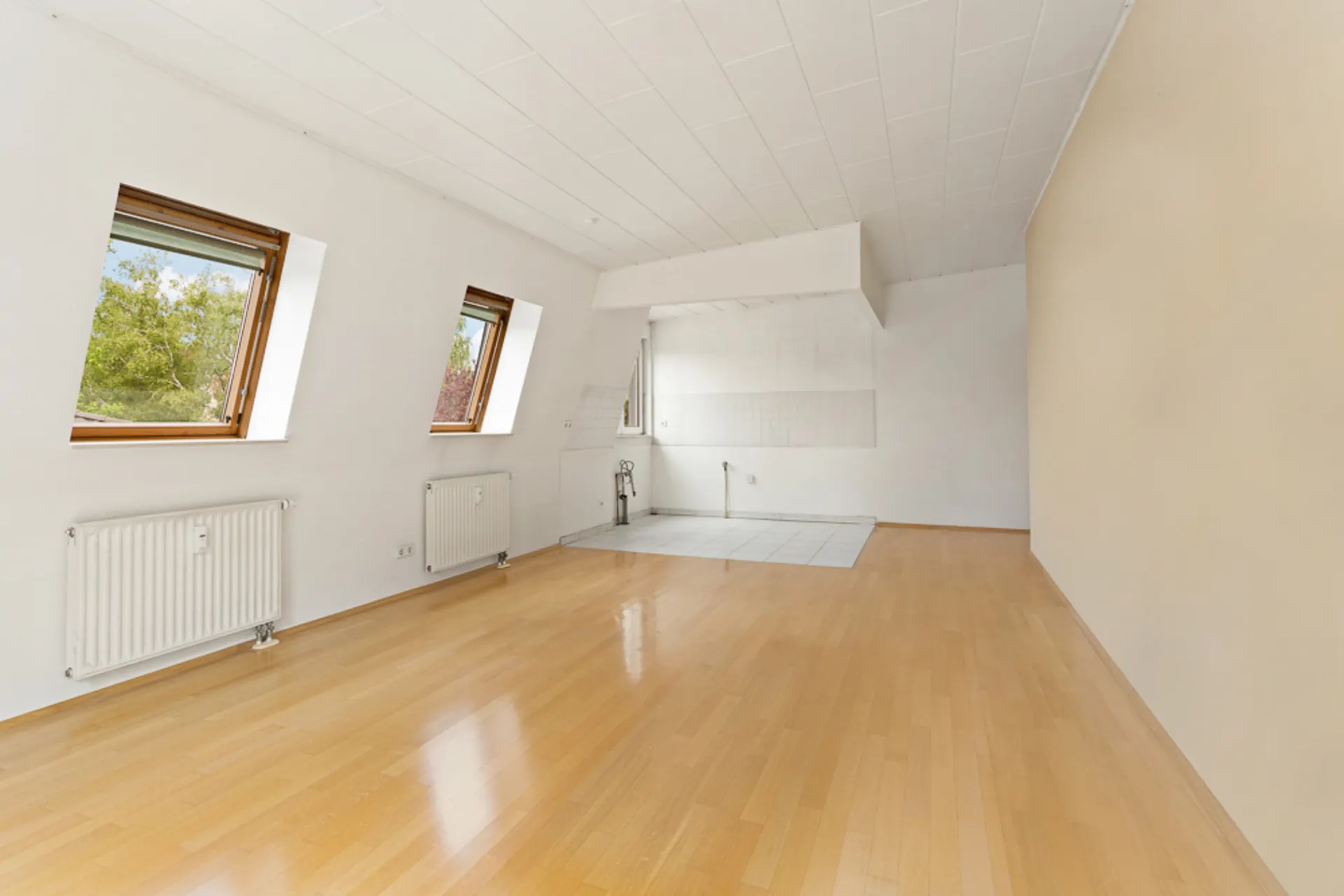 Leerer Raum mit zwei Fenstern, Holzboden und gefliestem Bereich für eine Küche.