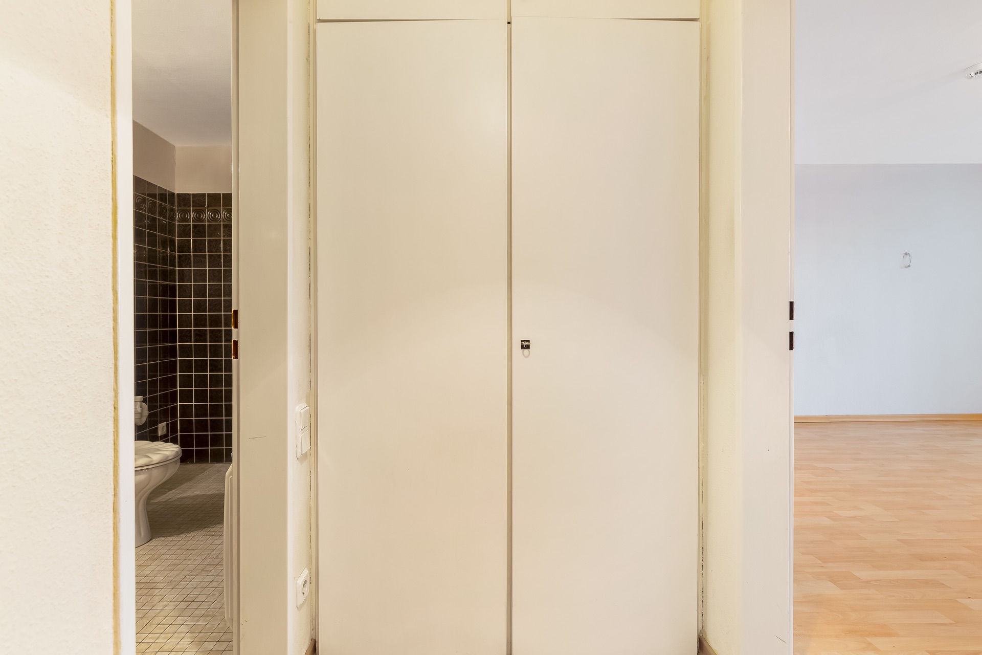 Flur mit weißem Einbauschrank, links ein Bad mit dunklen Fliesen, rechts ein Raum mit Holzfußboden.