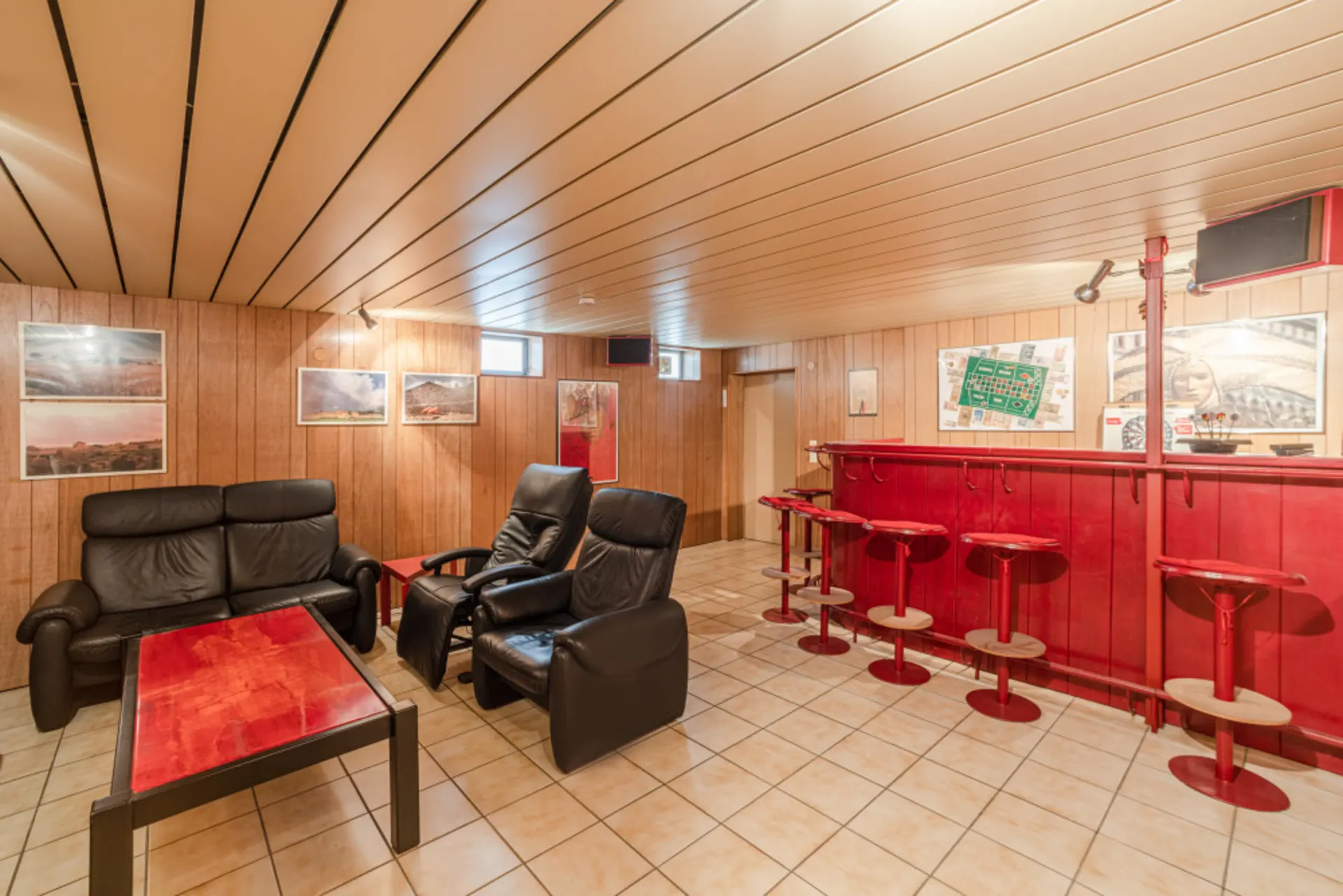 Wohnzimmer mit schwarzen Ledersesseln, rotem Tisch und einer roten Bar mit Barhockern.