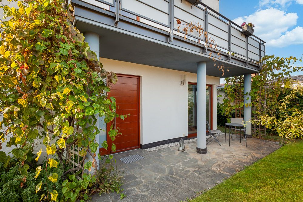 Überdachte Terrasse mit Tisch und zwei Stühlen neben einer roten Tür, umgeben von grüner Bepflanzung.