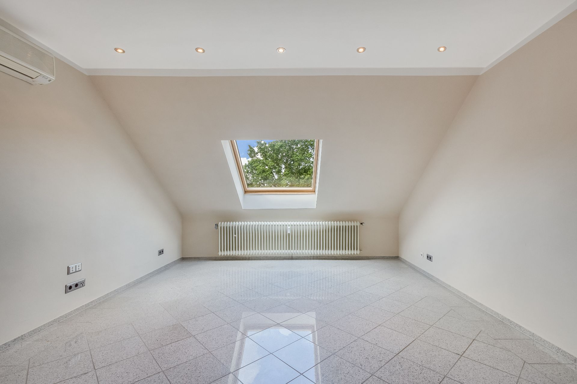 Leerer Raum mit Dachschräge, Fliesenboden, Dachfenster und Heizkörper unter dem Fenster.