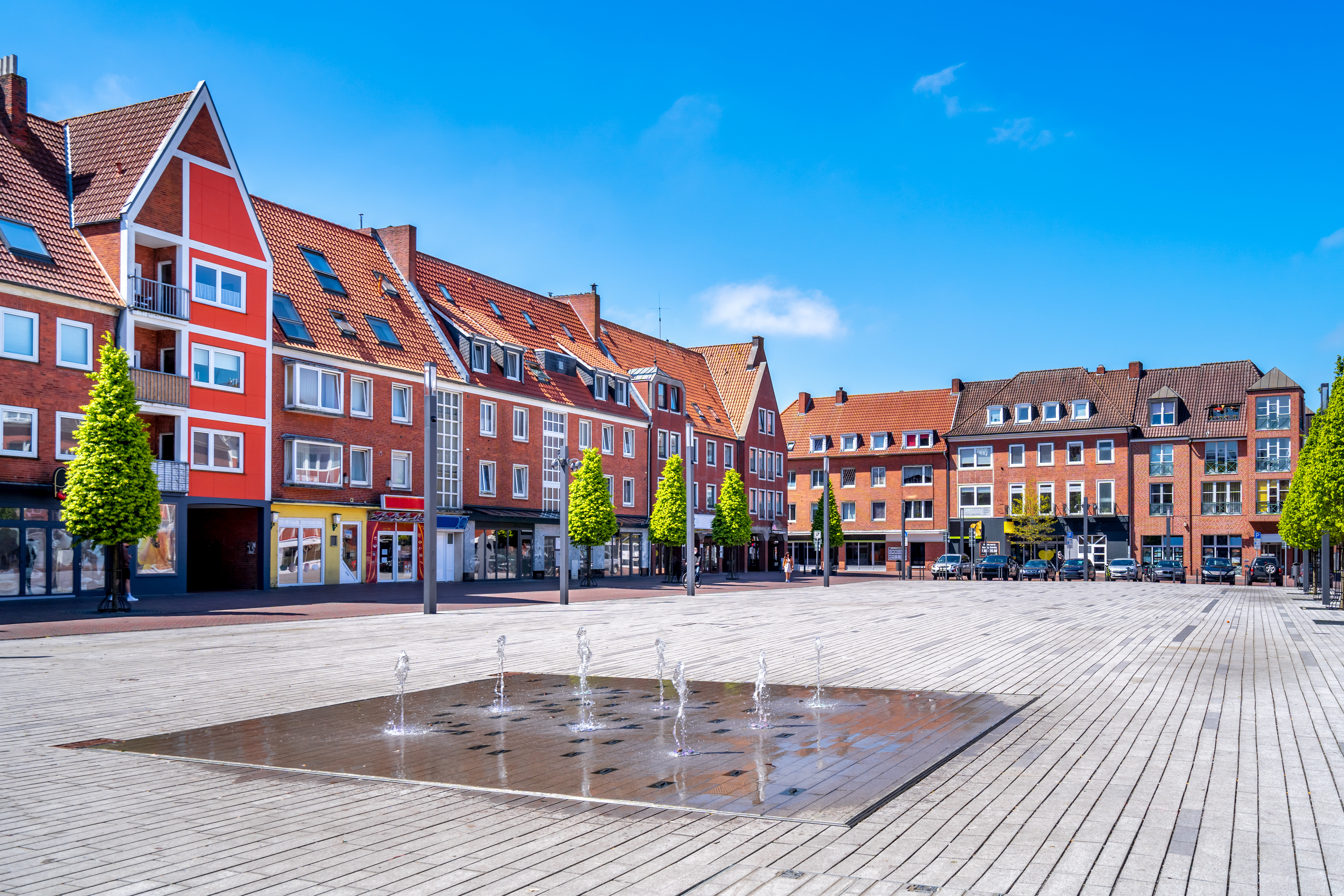 Moderner Platz mit Springbrunnen, umgeben von roten Wohnhäusern und Bäumen.