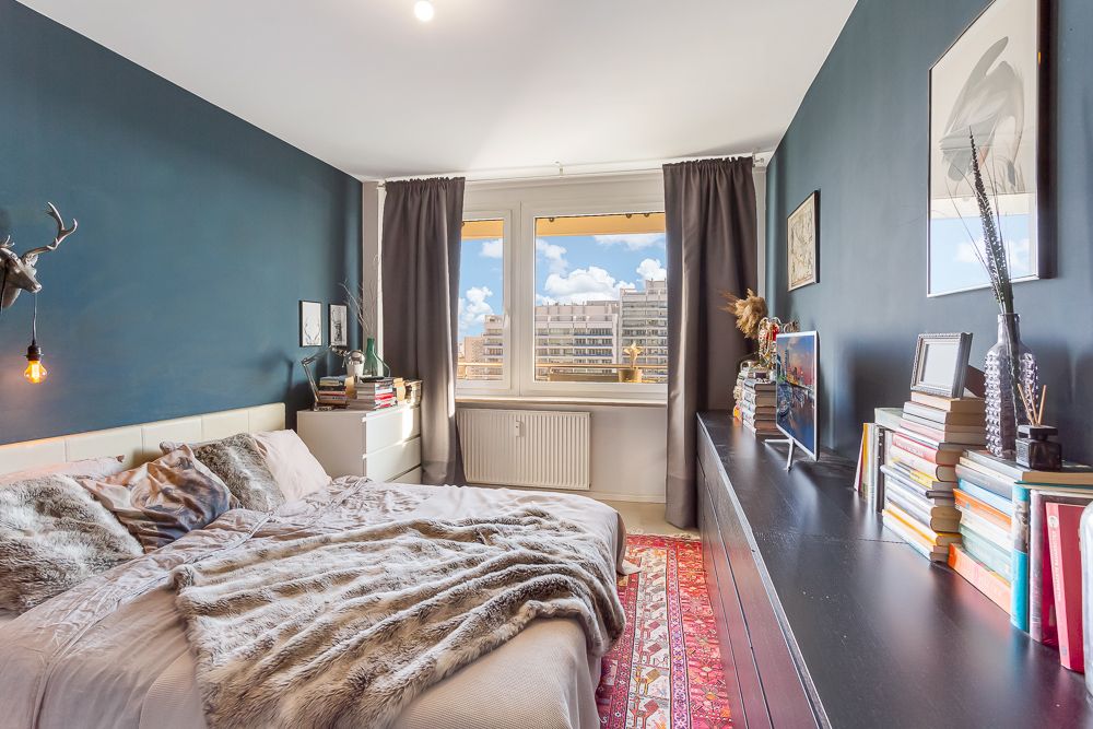 Modernes Schlafzimmer mit Doppelbett, Bücherregal, Teppich und großem Fenster mit Blick auf Nachbargebäude.