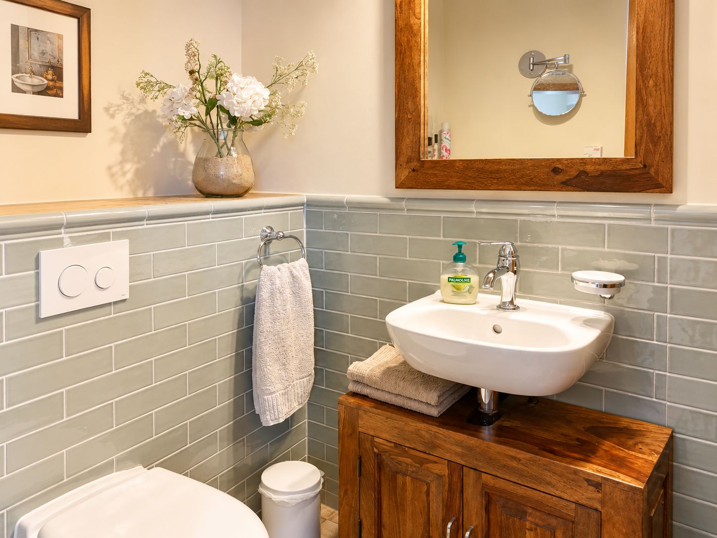 Kleines Bad mit Waschbecken, Spiegel, Toilette, Handtuch, Vase mit Blumen und hellen Fliesen.