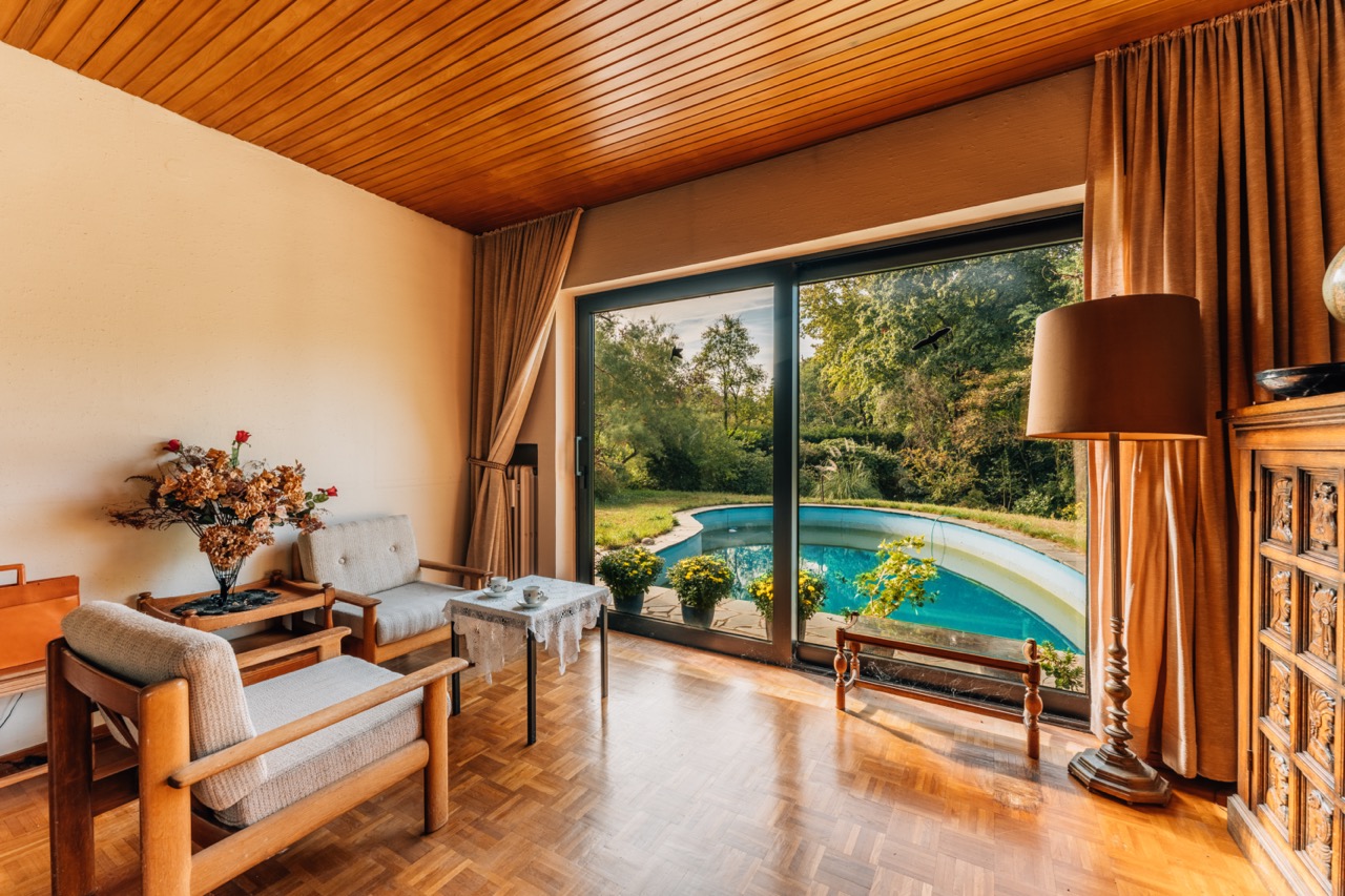 Wohnzimmer mit Holzmöbeln und Blick durch große Fenster auf einen Garten mit Pool.