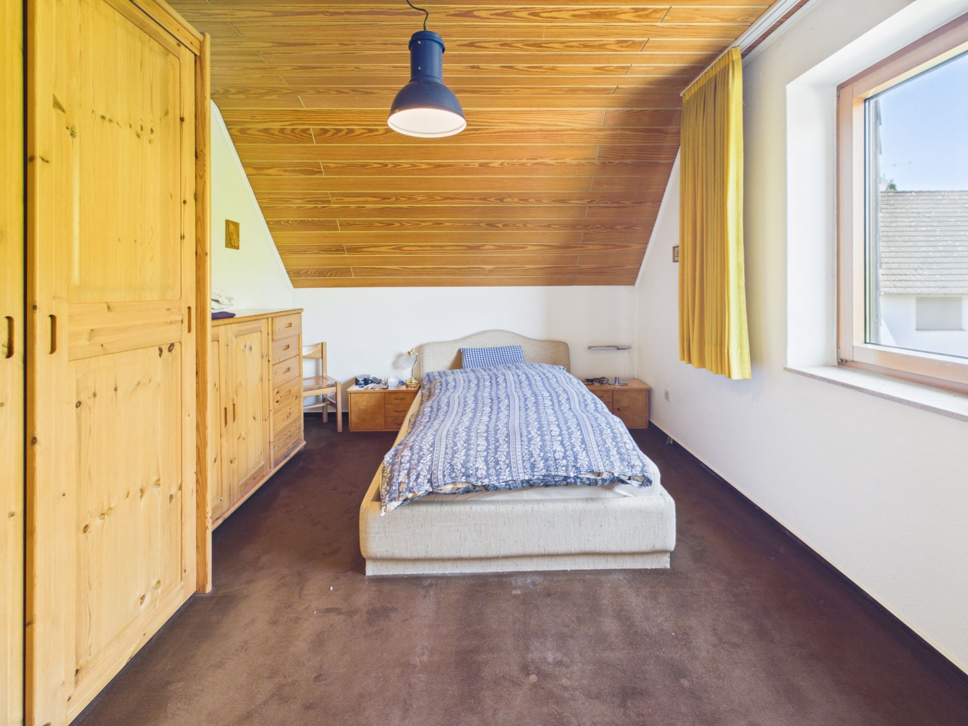 Schlafzimmer mit Einzelbett, Holzschrank, Kommode und Fenster unter schräger Holzdecke.
