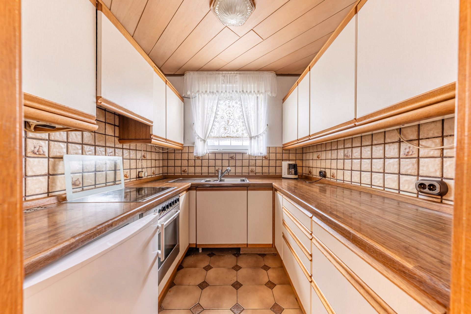 Schmale Küche mit Holzarbeitsflächen, Fliesenwand und Fenster mit Gardine.