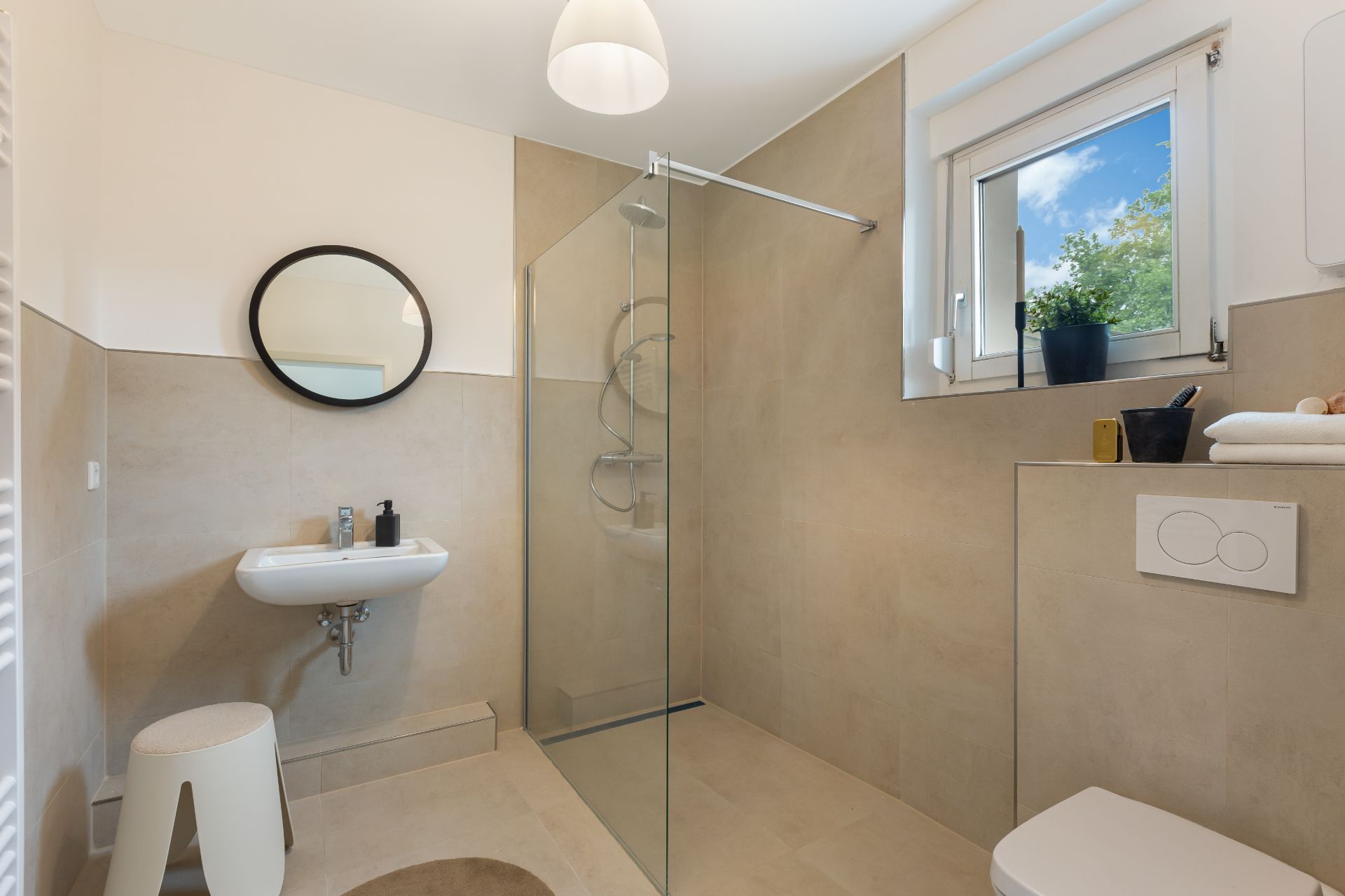 Modernes Badezimmer mit bodengleicher Dusche, Waschbecken, rundem Spiegel und Fenster.