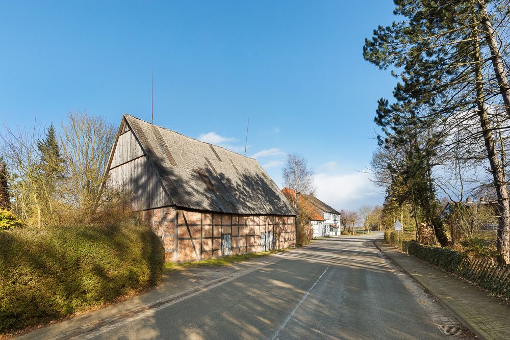 Fachwerkhaus mit großem Schrägdach an einer ruhigen Landstraße, umgeben von Bäumen.